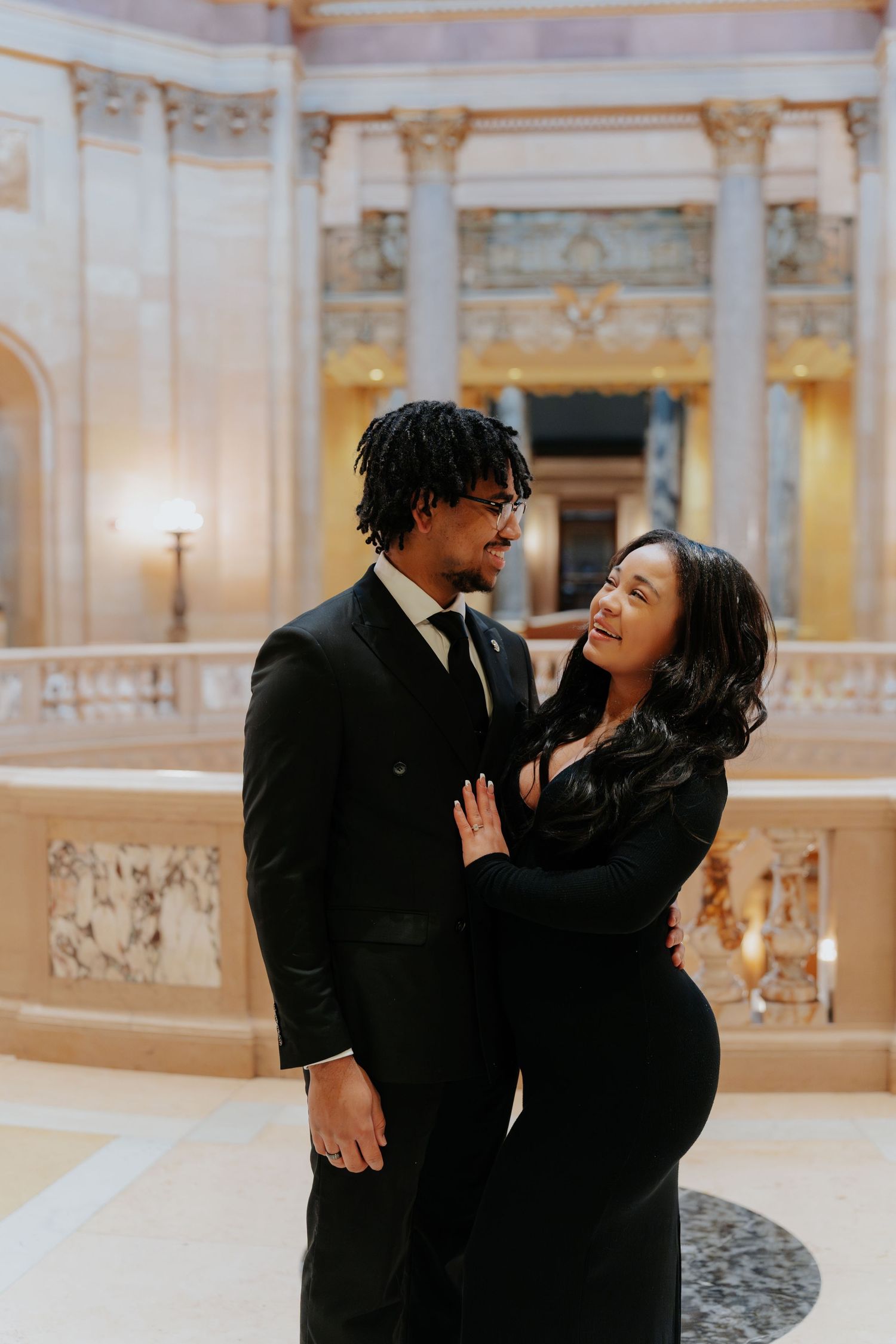Elegant couple in matching black attire sharing a romantic moment inside a grand ornate building.