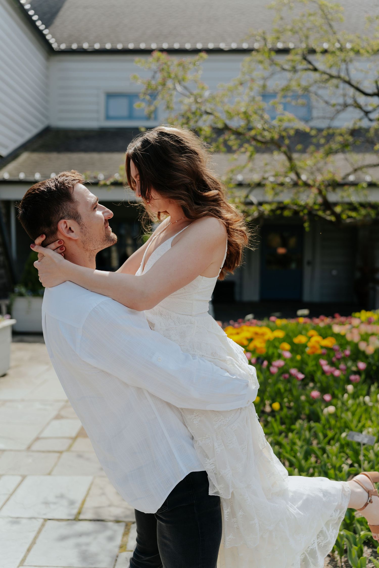 A groom lifts his bride joyfully outside a venue with colorful tulips blooming in the background.