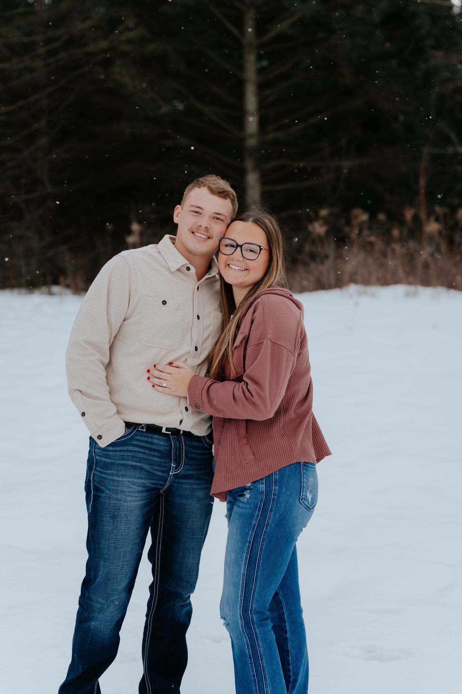 A smiling couple embraces in a snowy winter landscape, wearing casual jeans and sweatshirts with evergreen trees behind them.