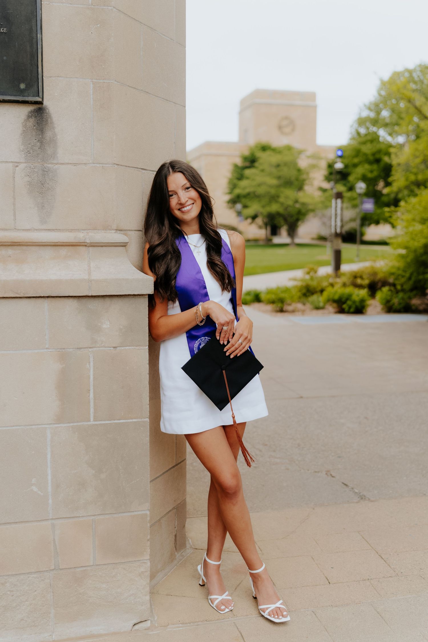 Smiling female graduate in purple top and white skirt holds graduation cap while leaning against stone column on campus.