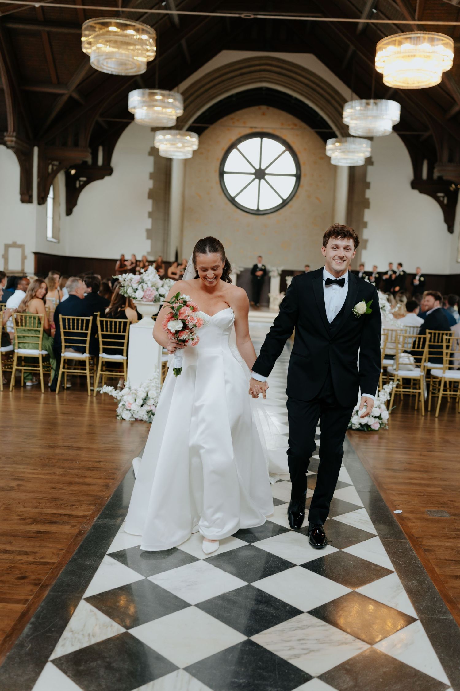 Newlyweds walk down checkered aisle in elegant hall, bride in white gown with bouquet, groom in black tuxedo.