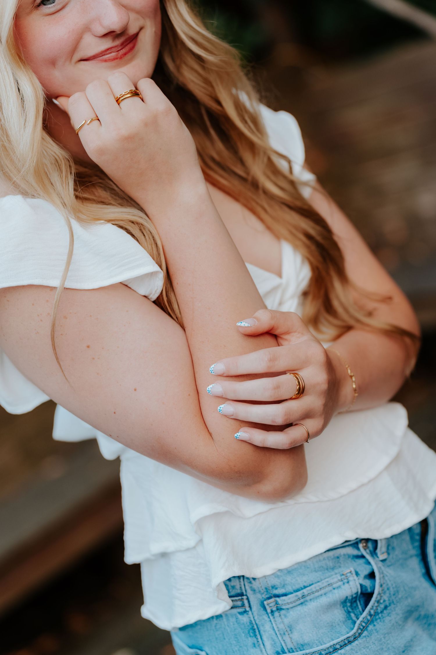 Blonde woman in white ruffled top and denim shorts wearing gold rings, posing with hand near face.