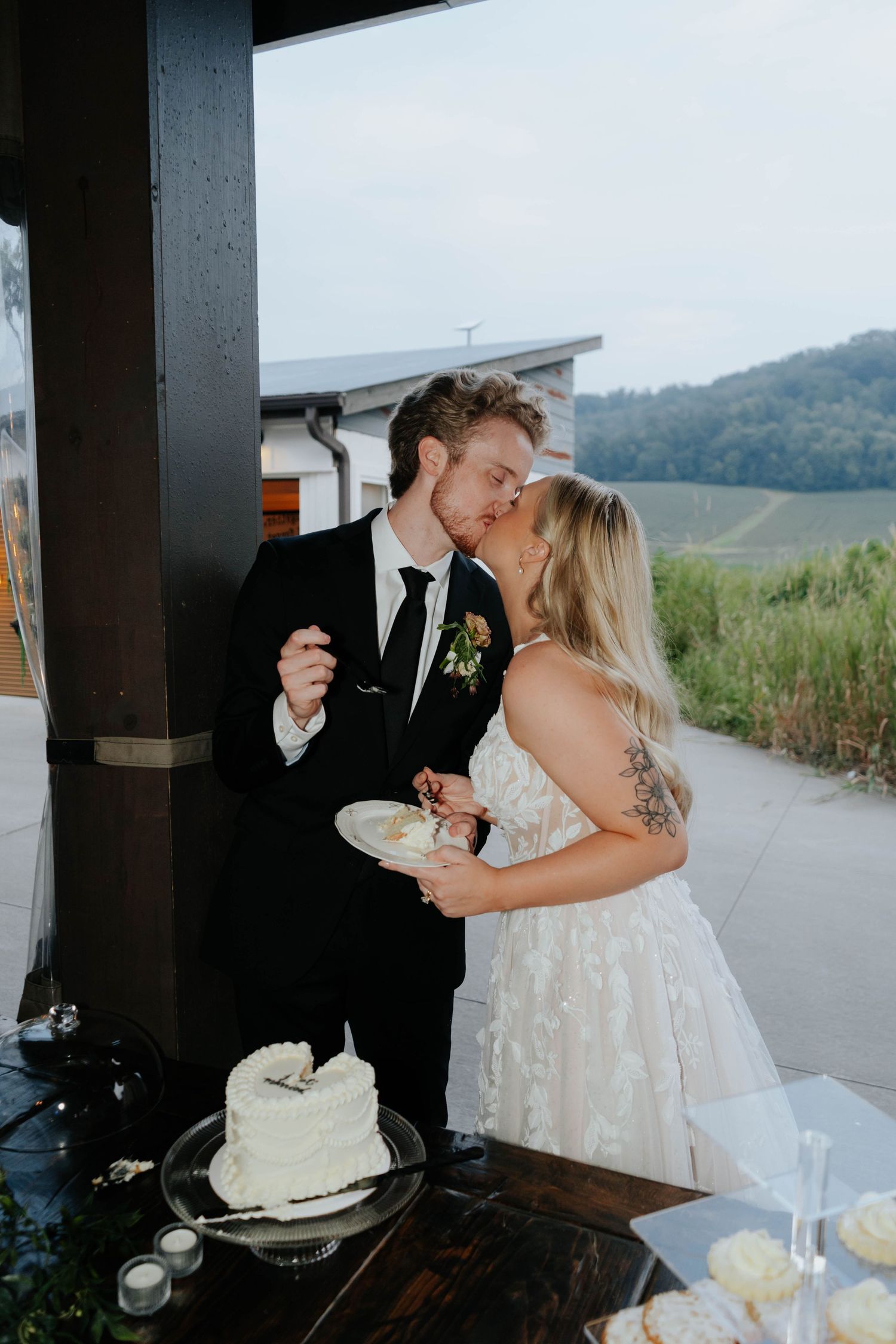 Bride and groom share a kiss while cutting their white wedding cake at an outdoor venue with scenic hills.