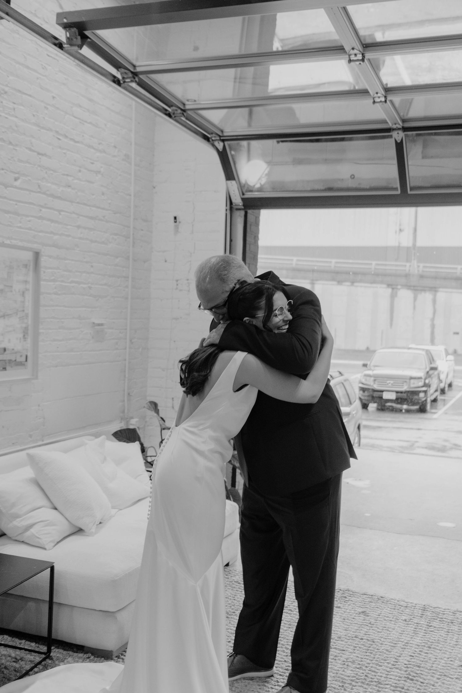 Black and white photo of a bride in a white gown sharing an emotional hug with a man in a suit at a wedding venue.