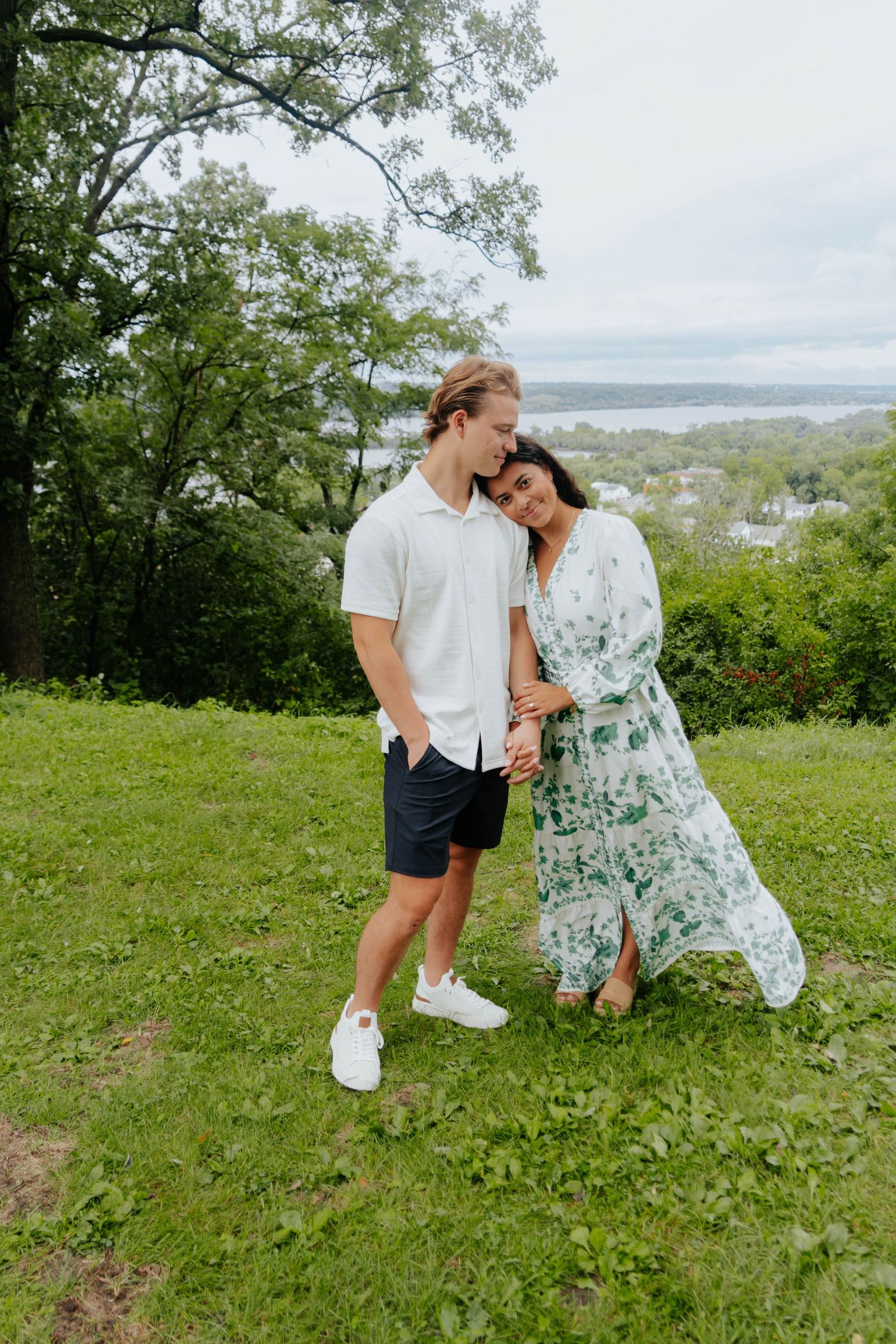 Couple embracing on a green hill, woman in floral dress, man in white shirt and navy shorts, with scenic water view.