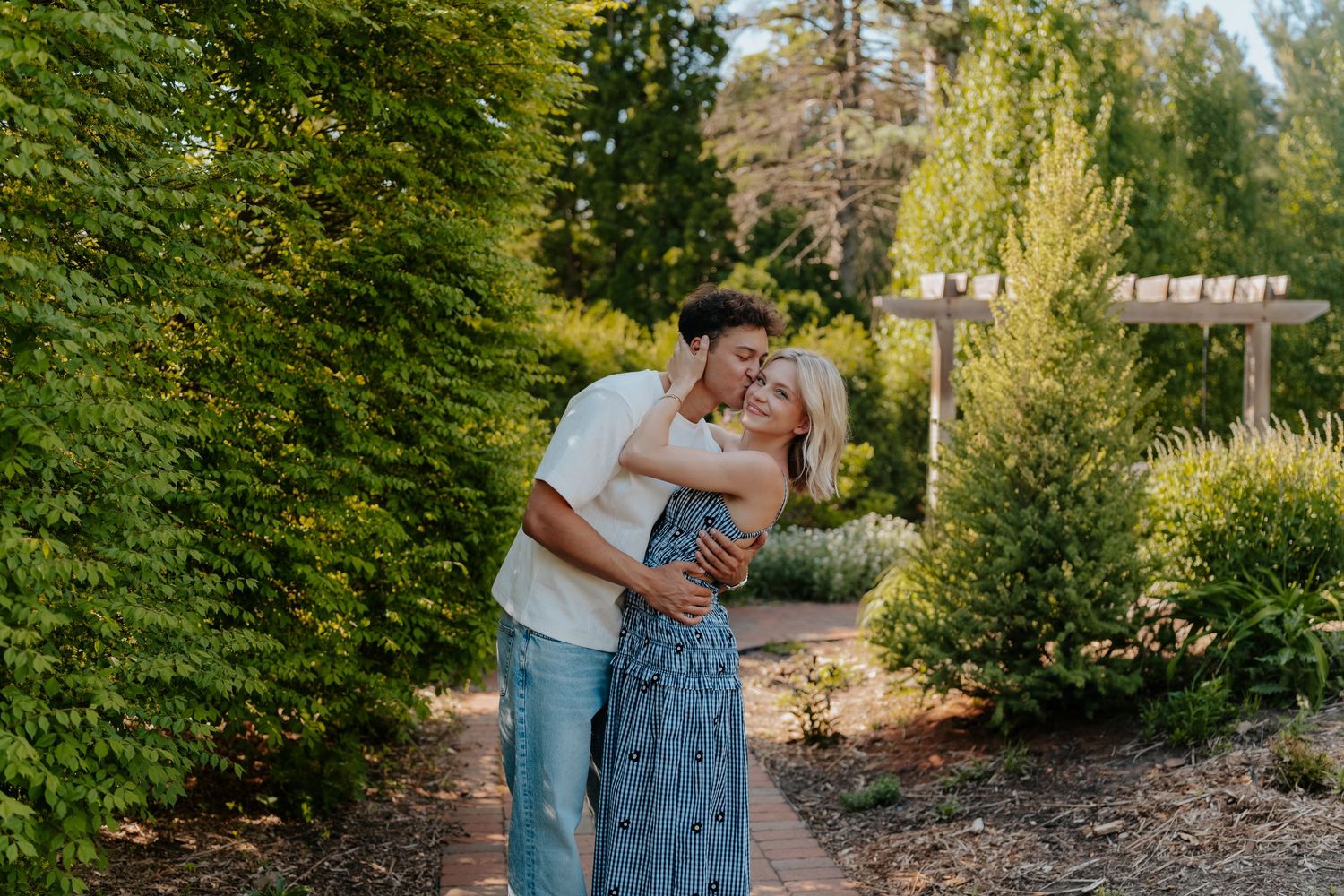 A couple embraces and kisses in a lush garden pathway surrounded by tall green trees and shrubs.