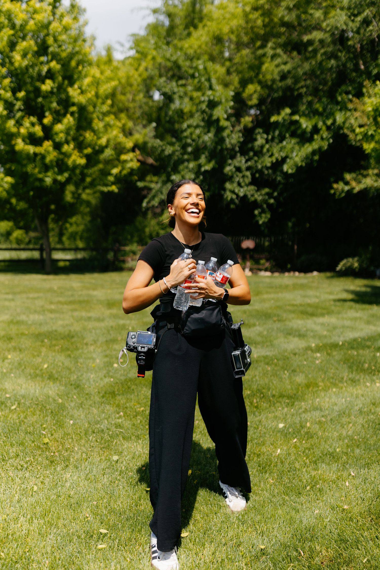 Happy woman in black outfit holding water bottles, laughing outdoors in a sunny green park.