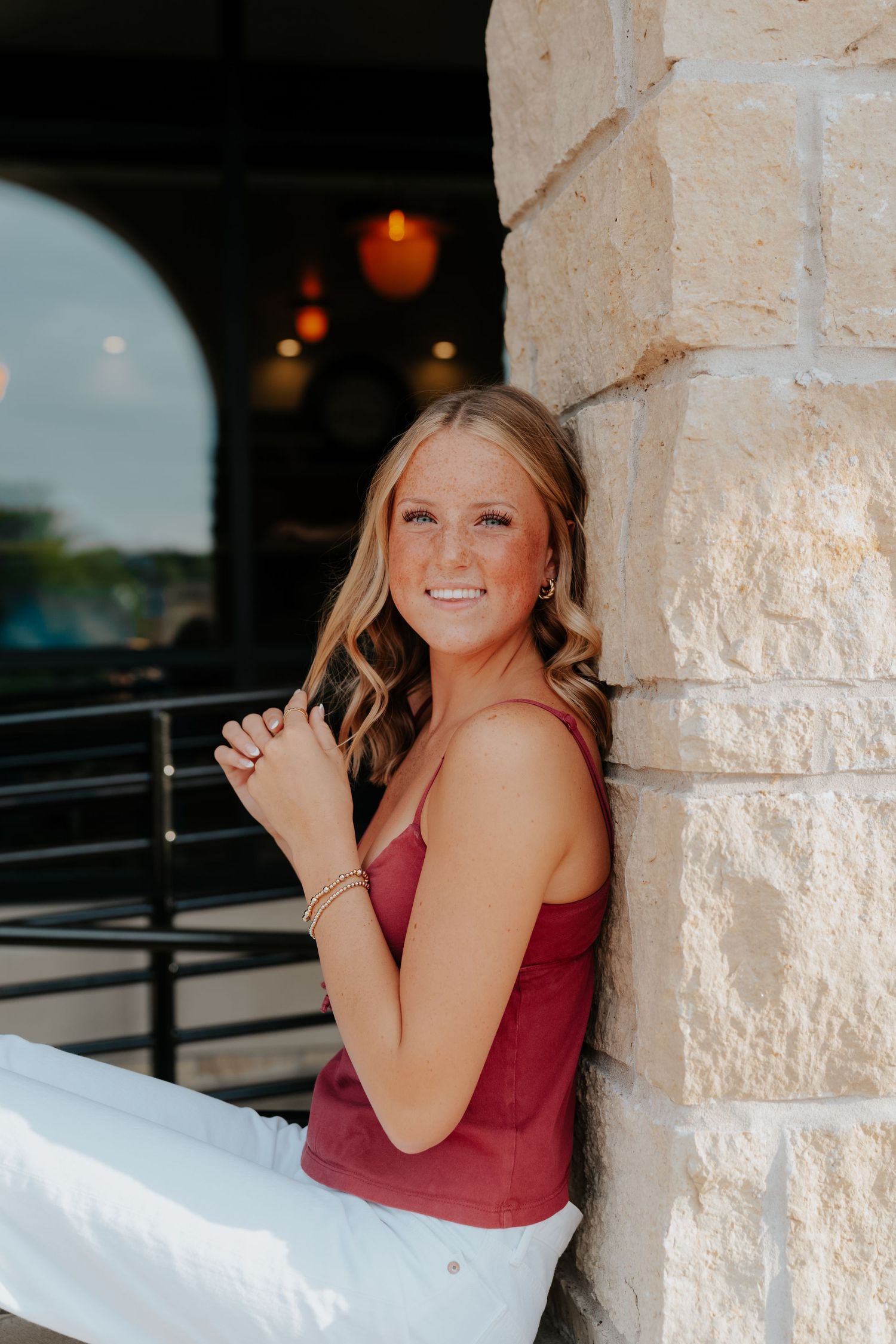 Young blonde woman in red tank top and white pants smiling while leaning against a stone pillar outdoors.