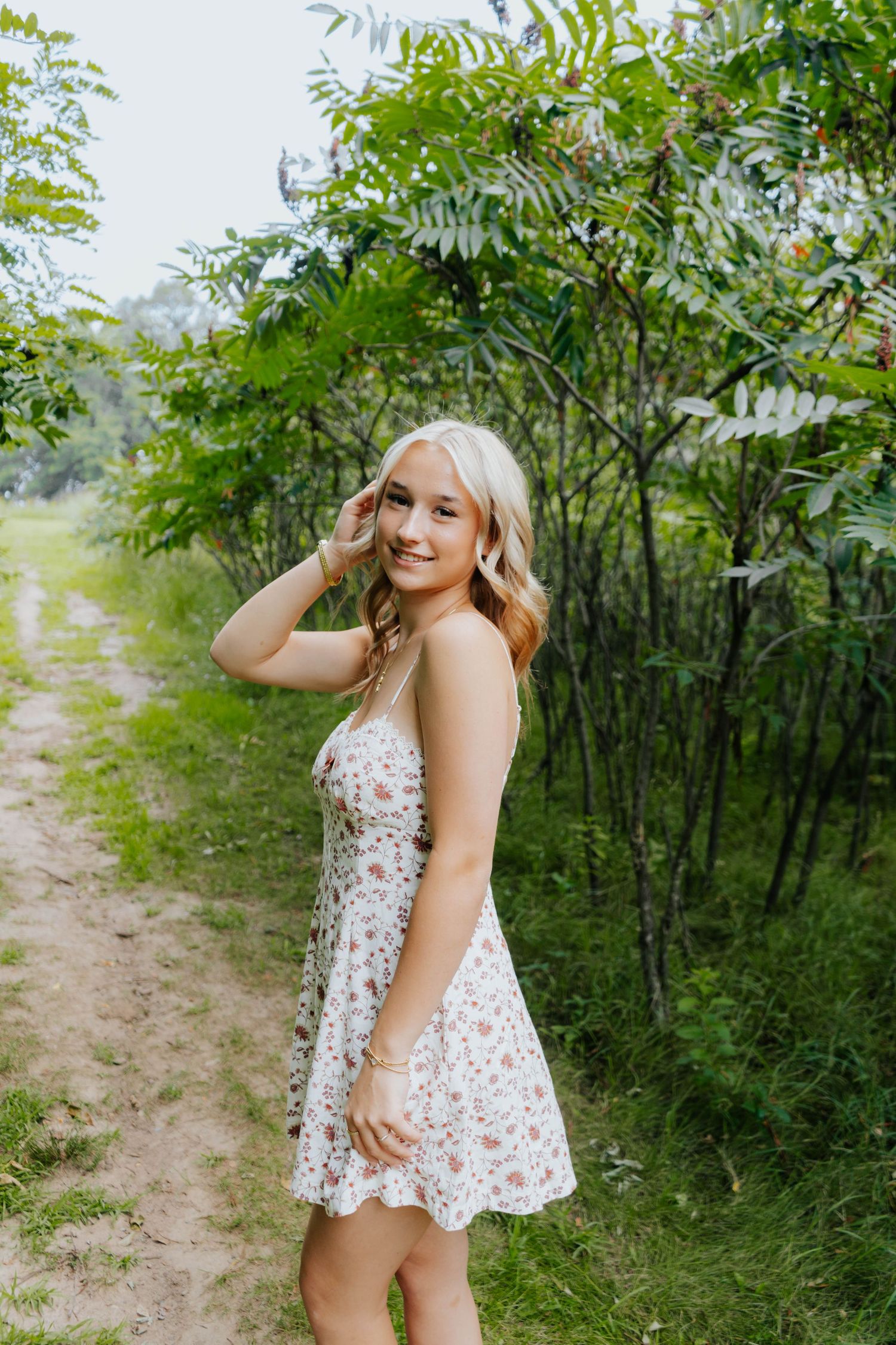 Young blonde woman in floral sundress smiling on a nature path surrounded by lush green trees.