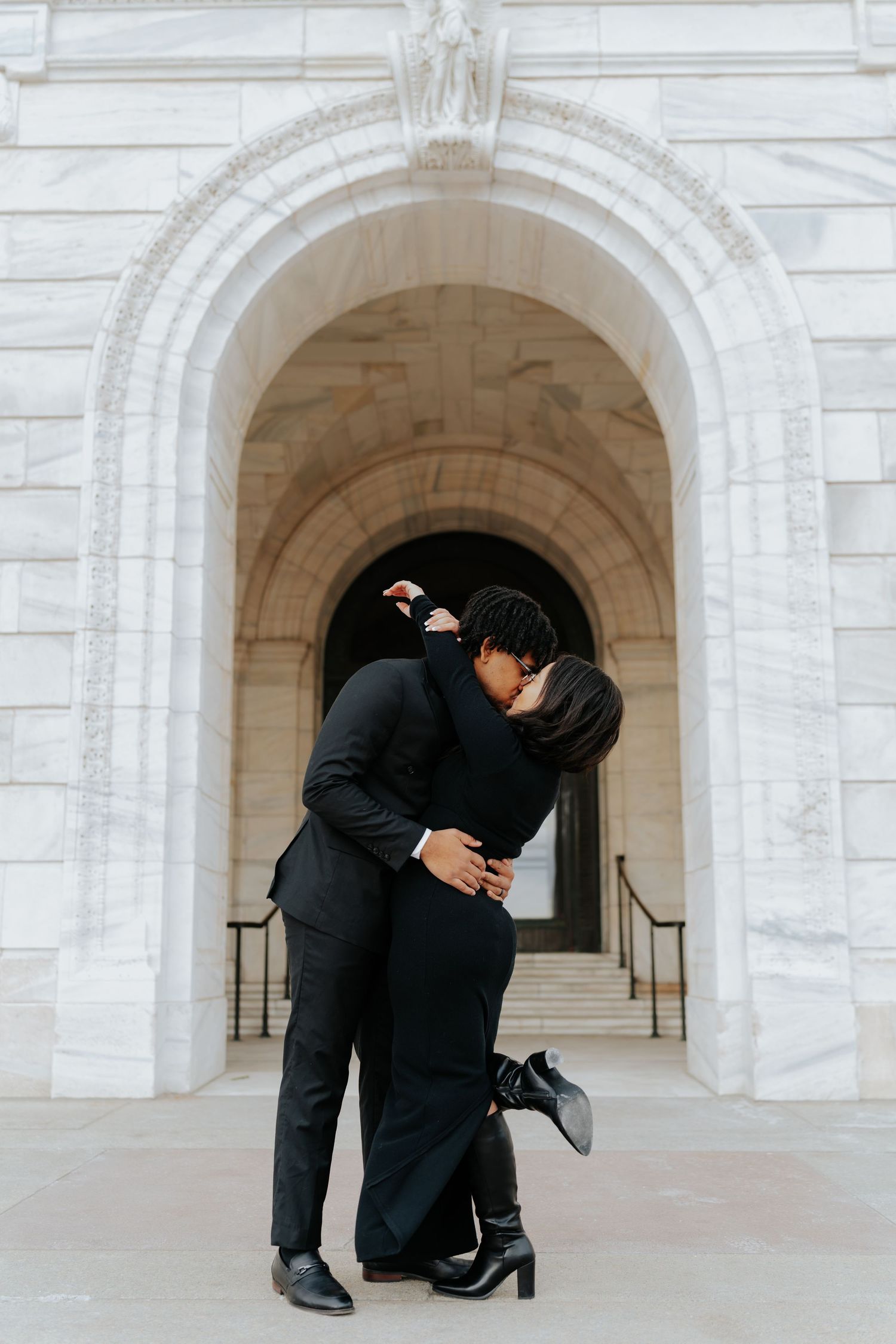 Couple in black attire sharing a romantic kiss under a grand marble archway of a classical building.