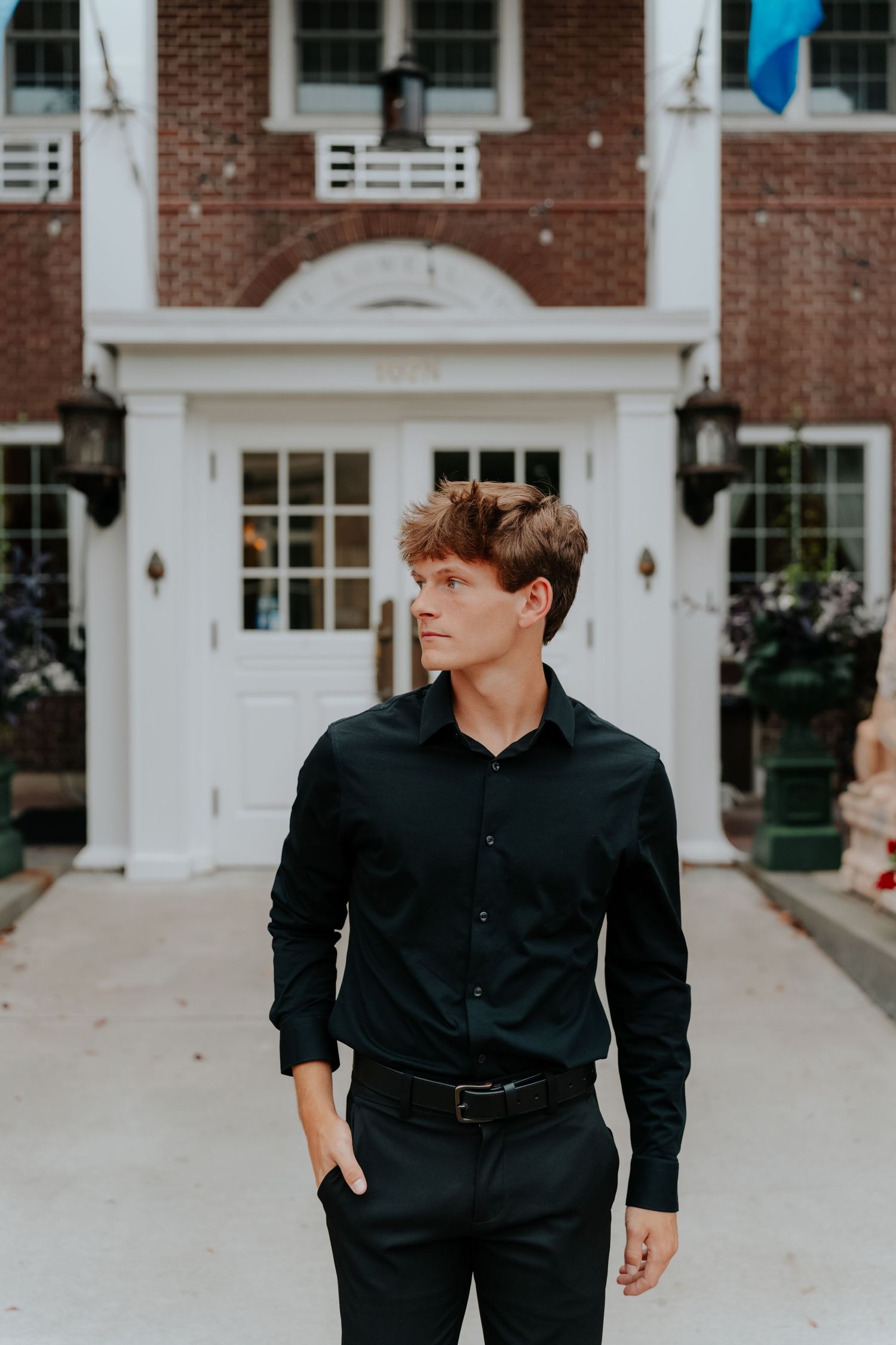Young man in black dress shirt and pants standing confidently in front of a brick building with white entrance.