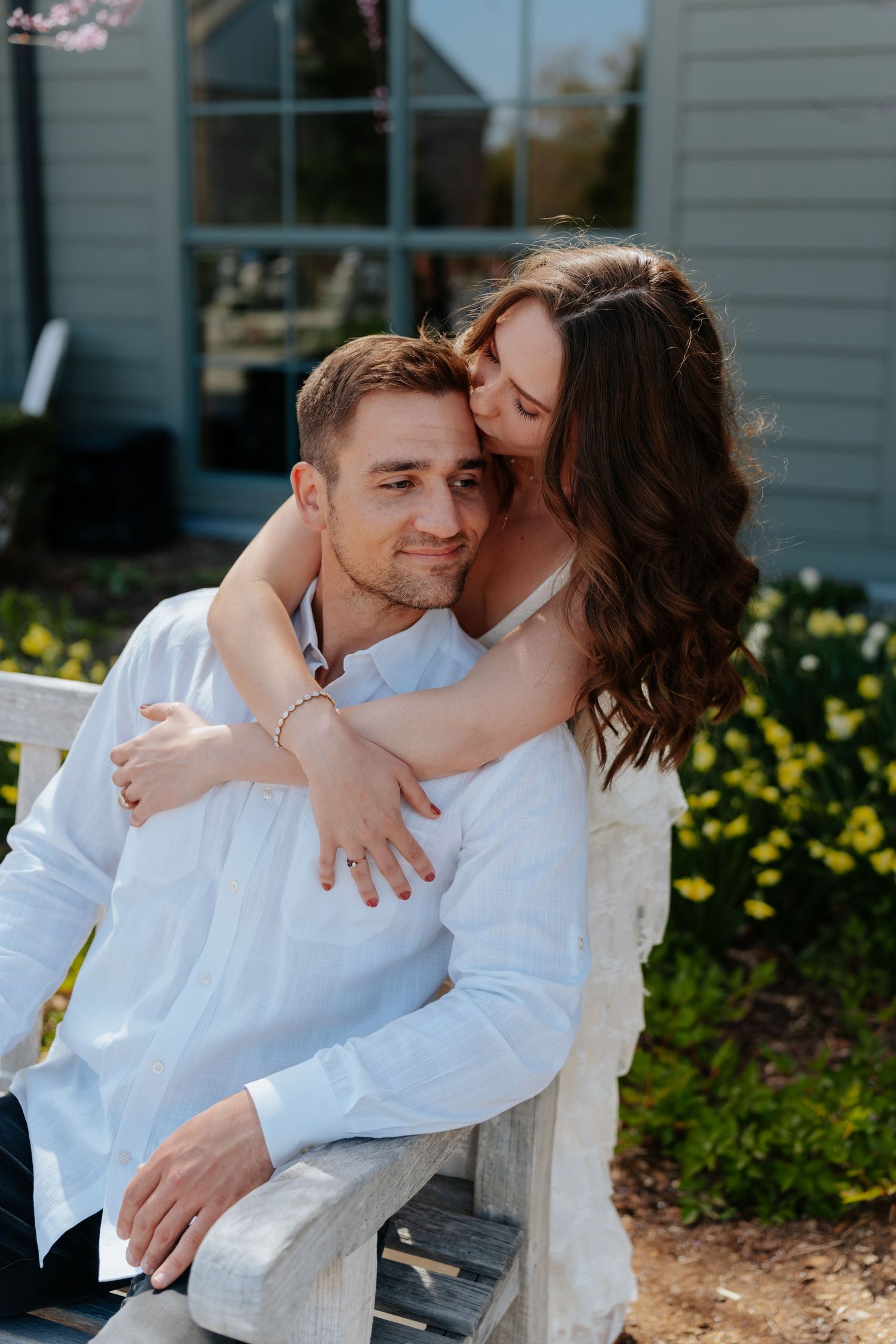 Couple in white outfits sharing a tender embrace on a garden bench with yellow flowers in the background.