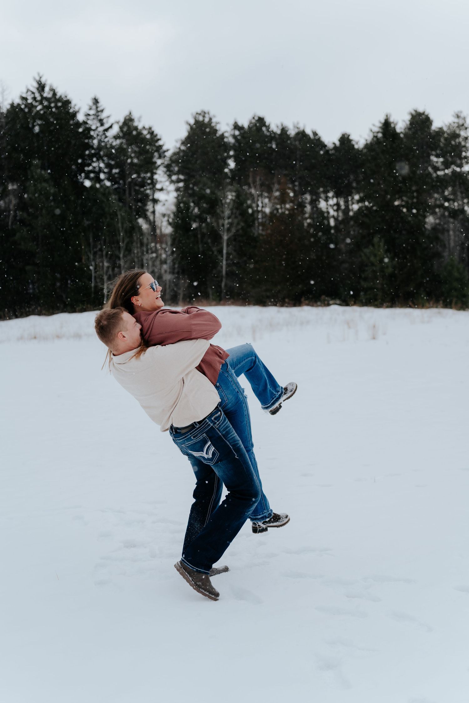 Couple sharing a romantic moment in snowy winter landscape, man lifting woman near evergreen forest.