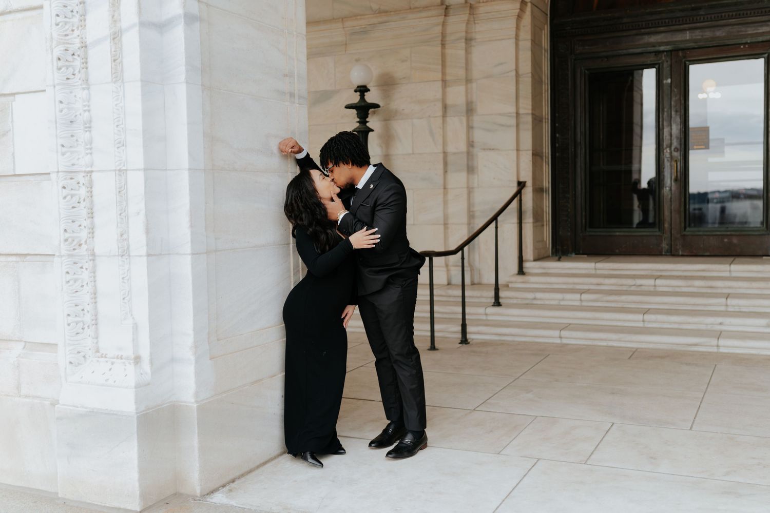 Couple in matching black outfits sharing a romantic kiss against a marble column at an elegant building exterior.