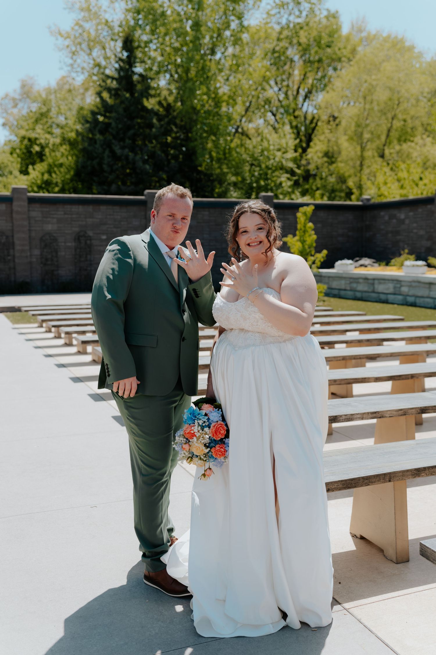 A newlywed couple in a green suit and white wedding gown show off their wedding rings at an outdoor venue.