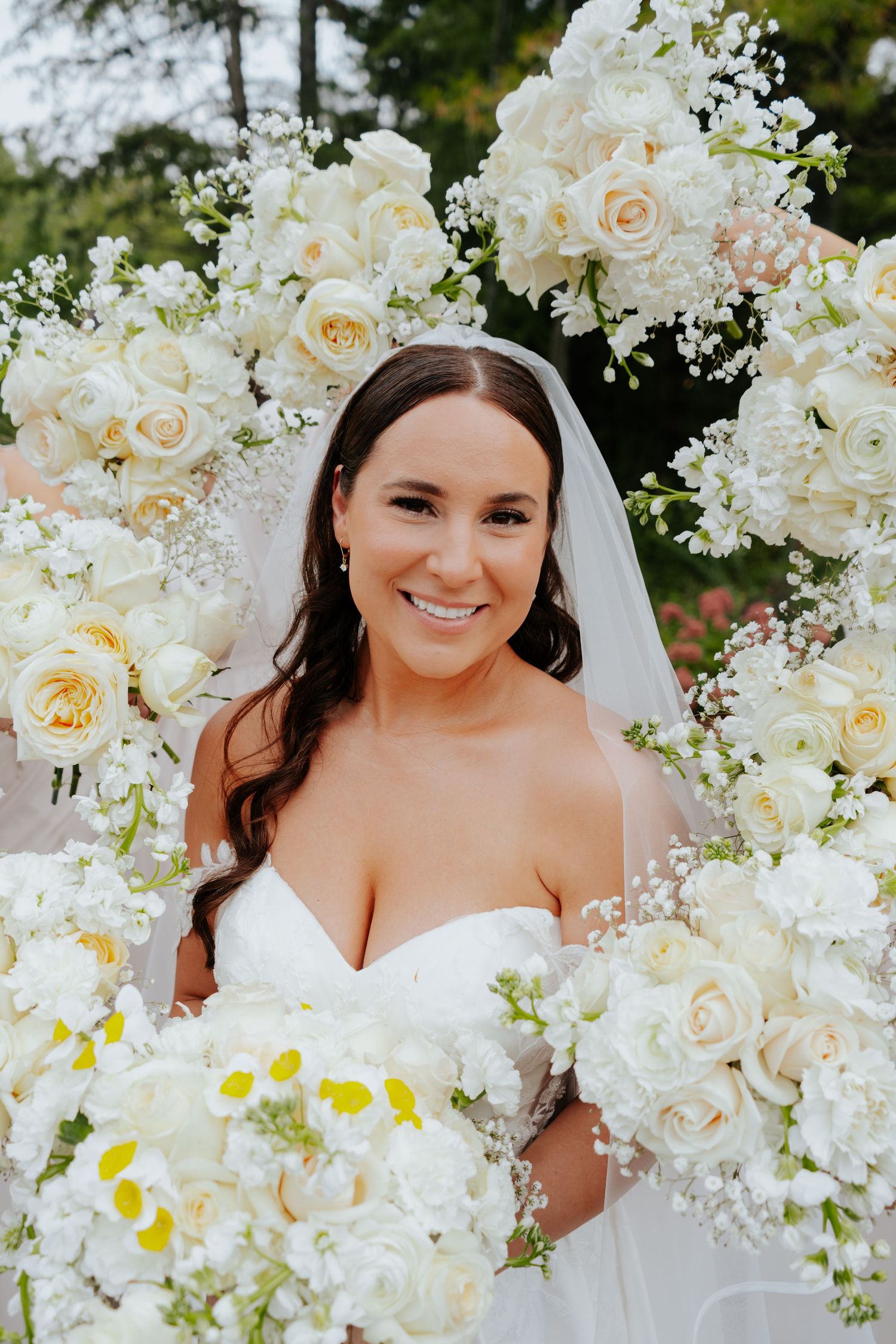 Smiling bride in white strapless gown and veil surrounded by lush white rose floral arch on her wedding day.