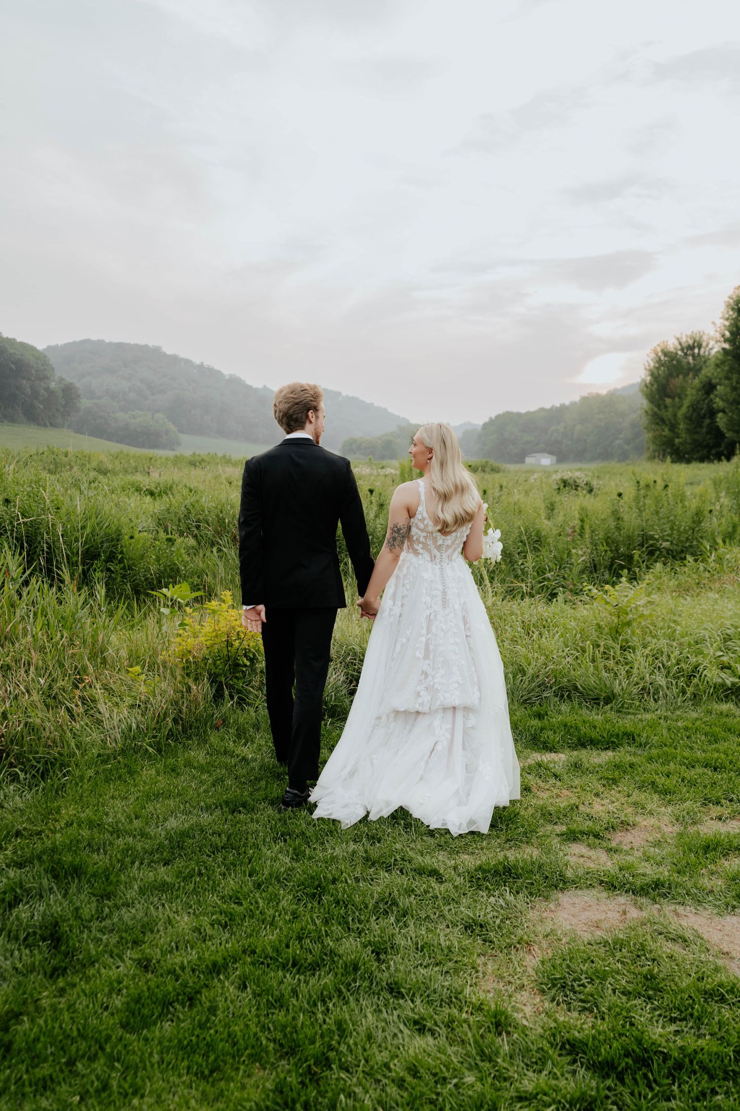 A bride and groom walk hand-in-hand through a lush green meadow, with rolling hills and a cloudy sky in the background.