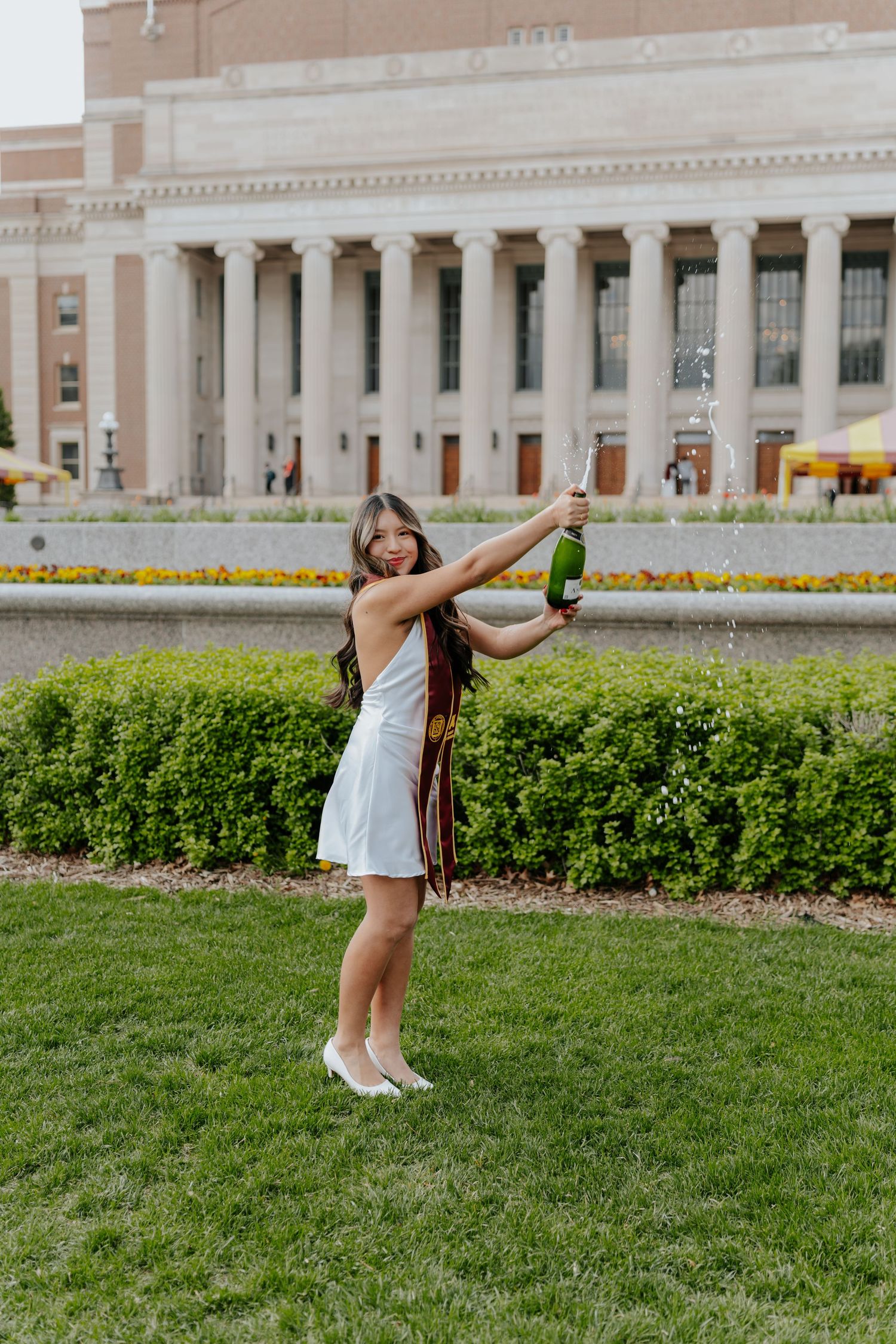 Woman in white dress popping champagne bottle on green lawn outside grand neoclassical building during graduation celebration.
