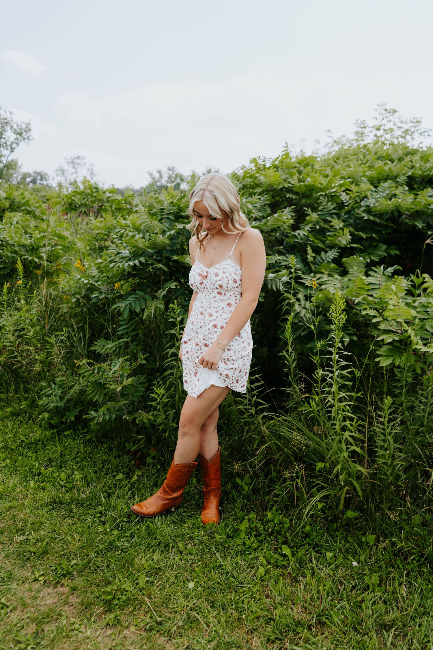 Blonde woman in white floral mini dress and brown cowboy boots standing in front of lush green bushes outdoors.