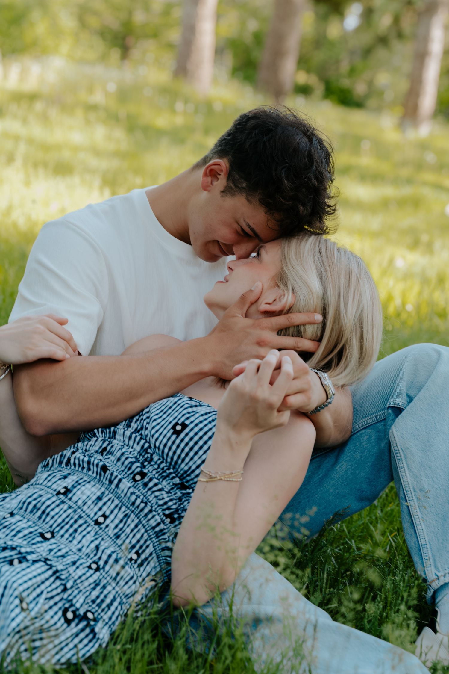 A couple sits on green grass, sharing a tender moment as they lean close together in a sunny park.