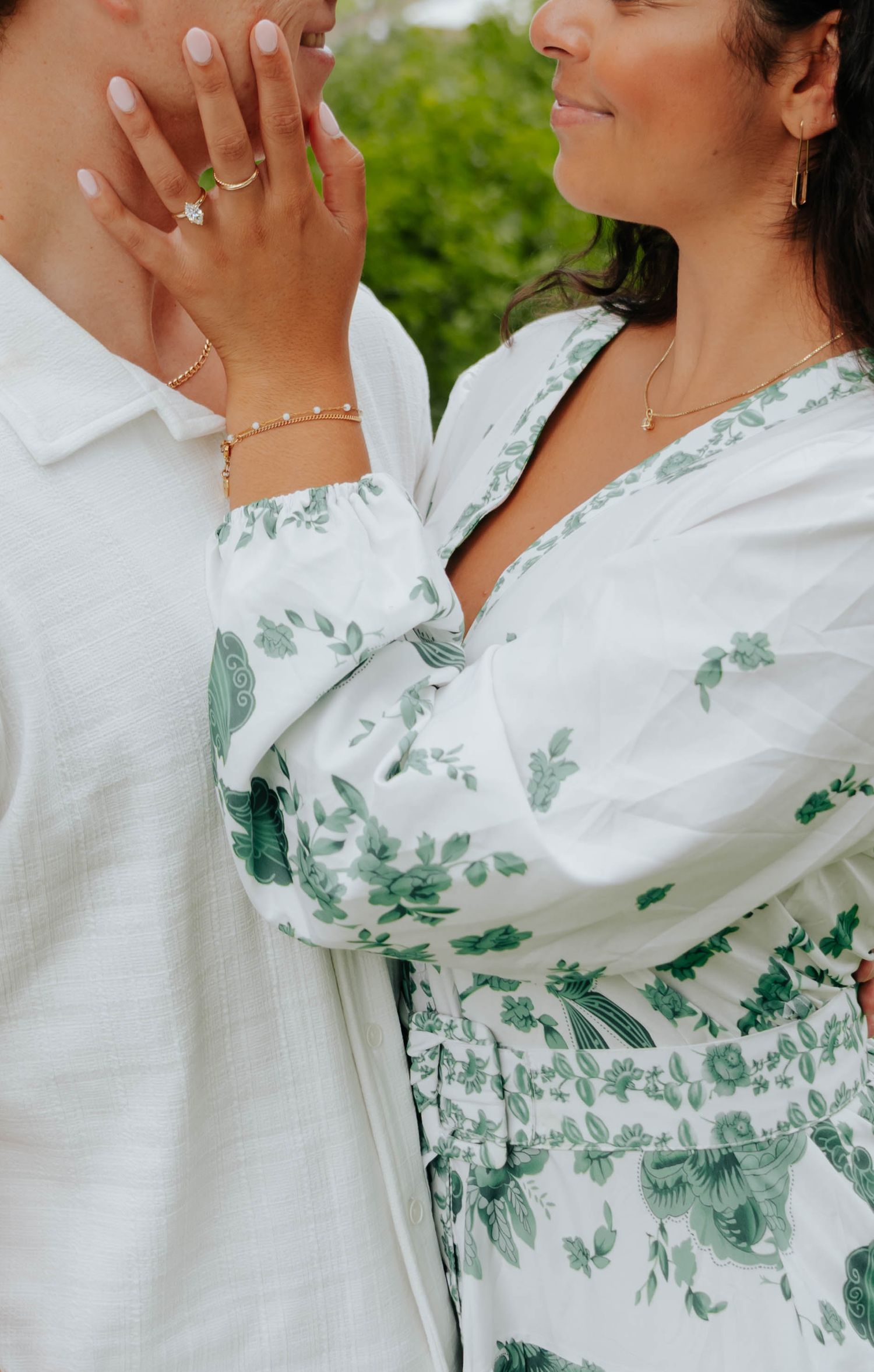 Couple in white and green floral outfits, woman wearing diamond ring and gold bracelet, embracing outdoors.