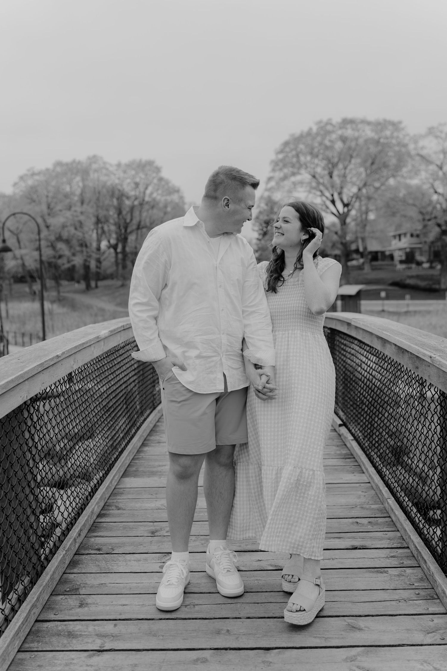 Black and white photo of a couple sharing a tender moment on a wooden bridge surrounded by trees.