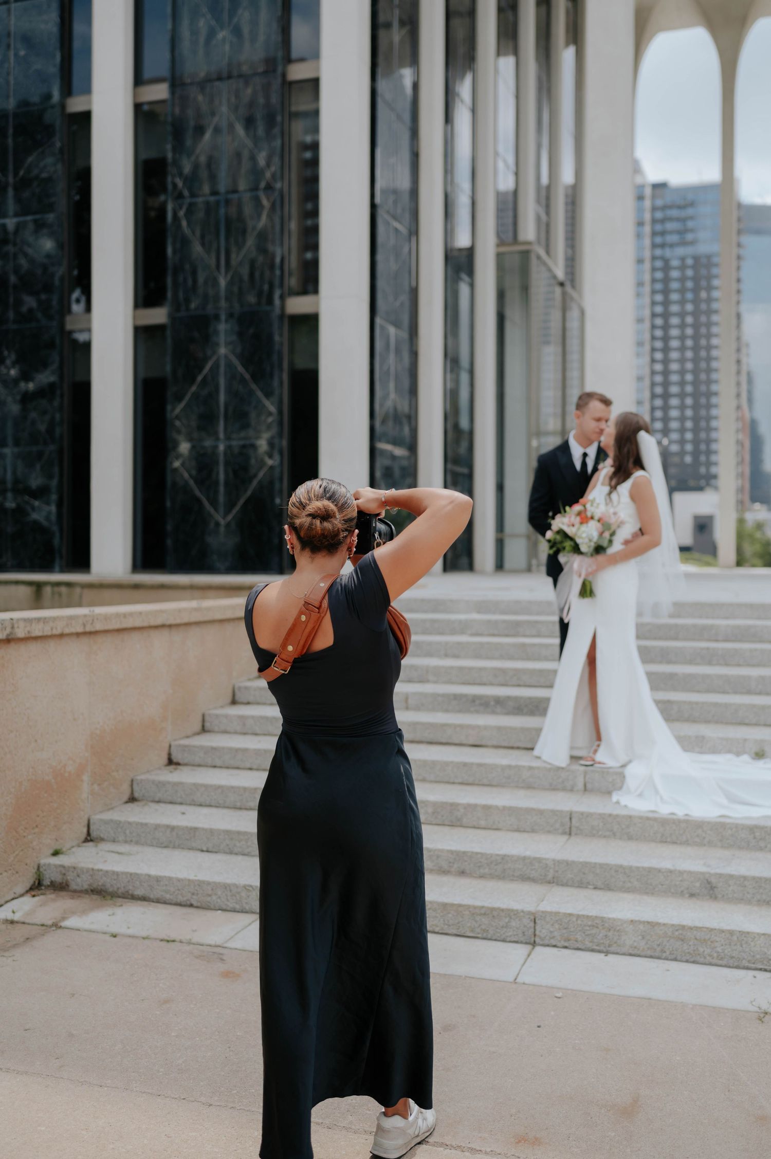 A photographer in black dress captures a wedding moment on elegant stone steps with city buildings in background.