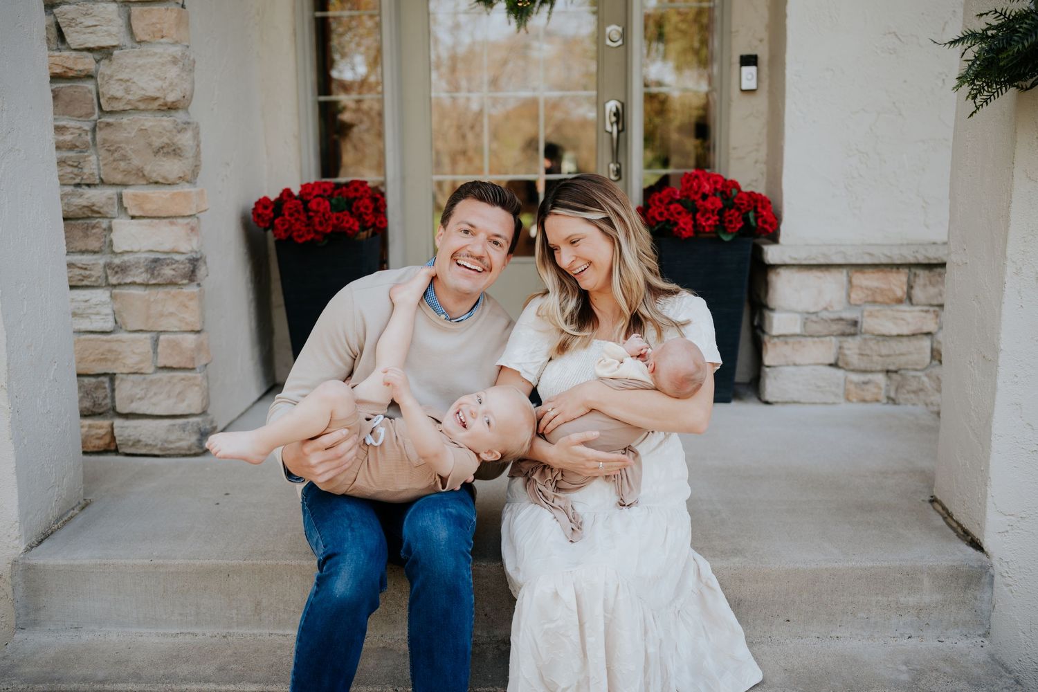 Happy couple sitting on front porch steps holding newborn twins, surrounded by red floral arrangements.