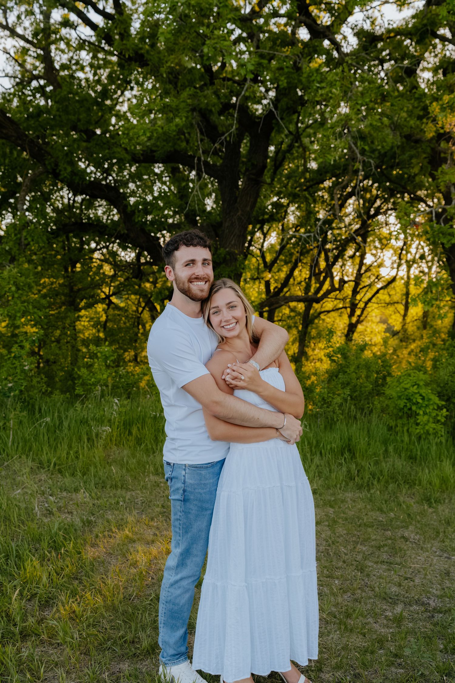 Happy couple hugging in lush green field at golden hour during outdoor engagement photography session.