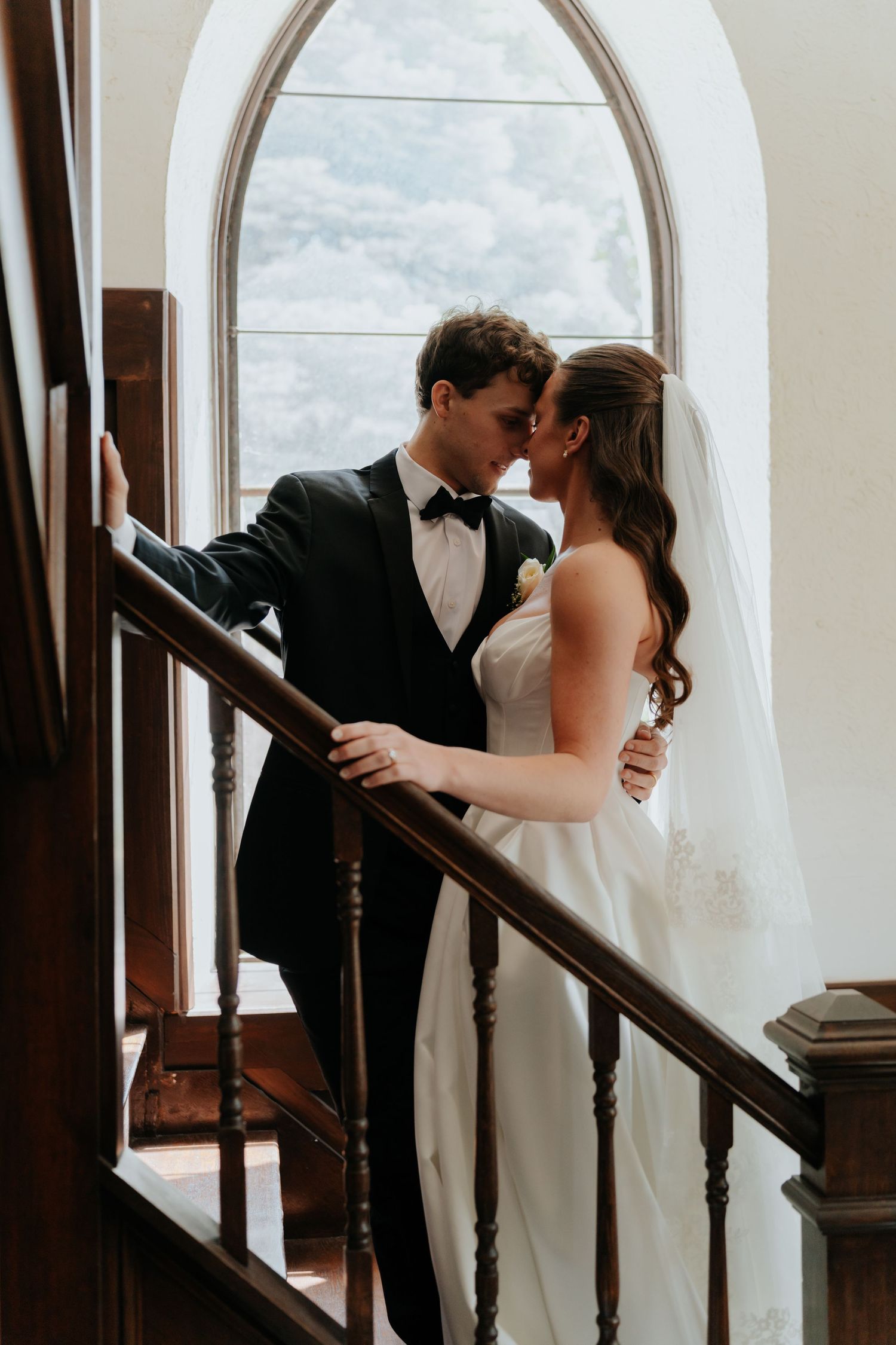 Bride and groom share a romantic moment on a staircase by an arched window, dressed in wedding attire.