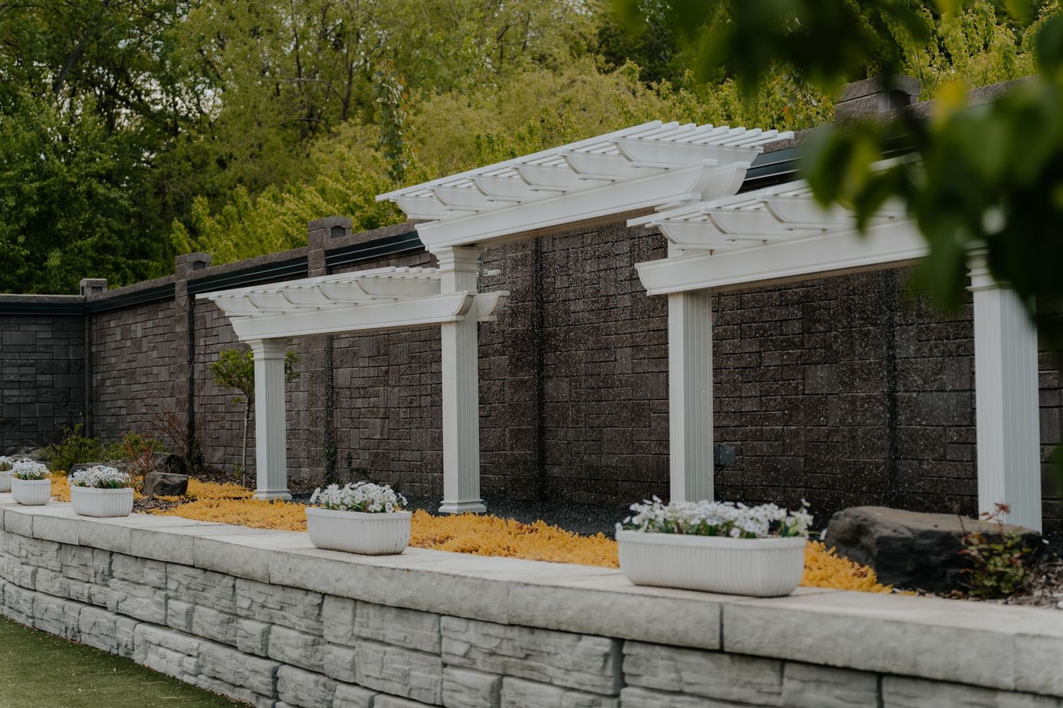 White pergola columns against a dark stone wall with planter boxes and mulch landscaping in an outdoor garden setting.