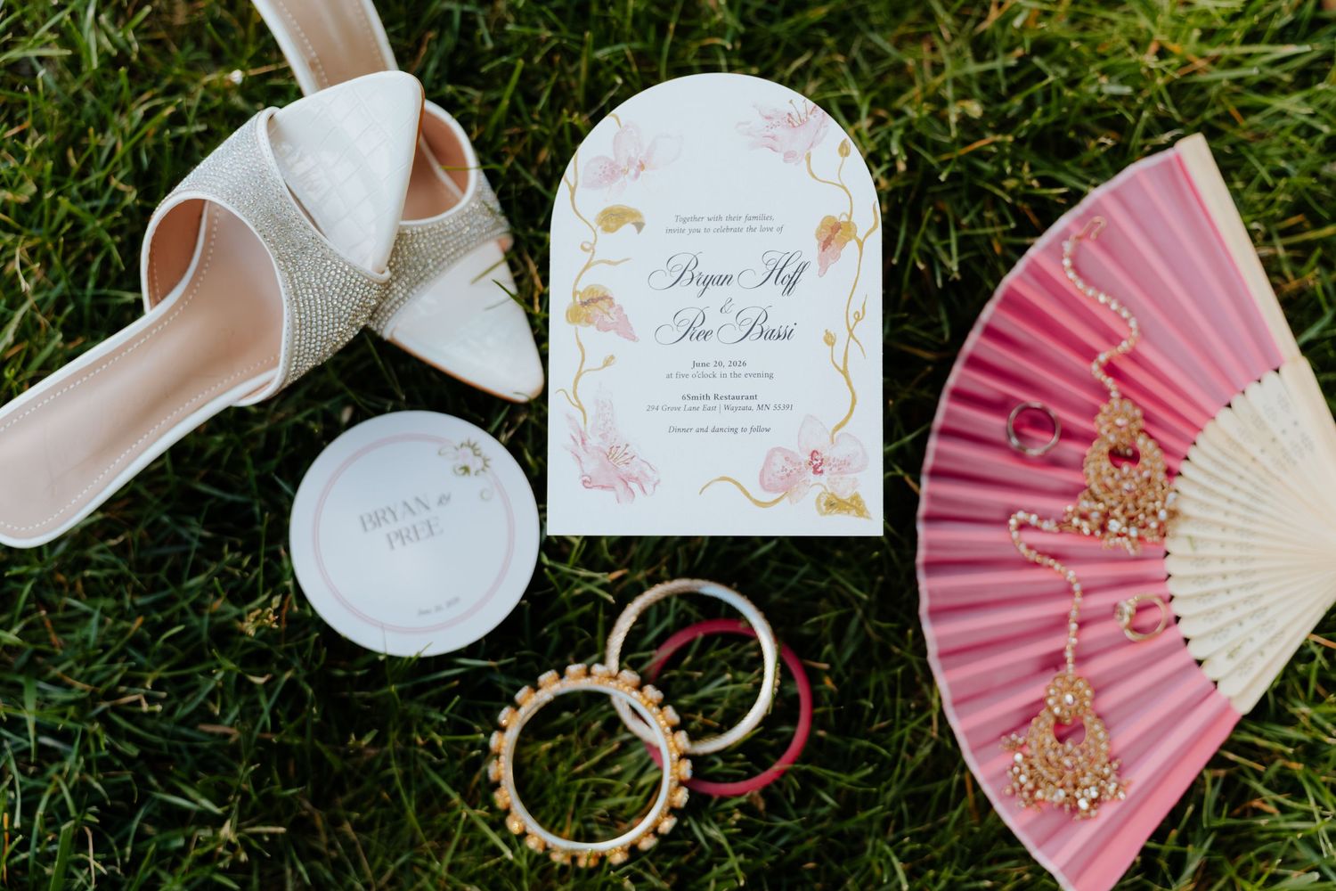 Wedding flat lay featuring white heels, pink fan, gold earrings, bangles, invitation, and compact on green grass.