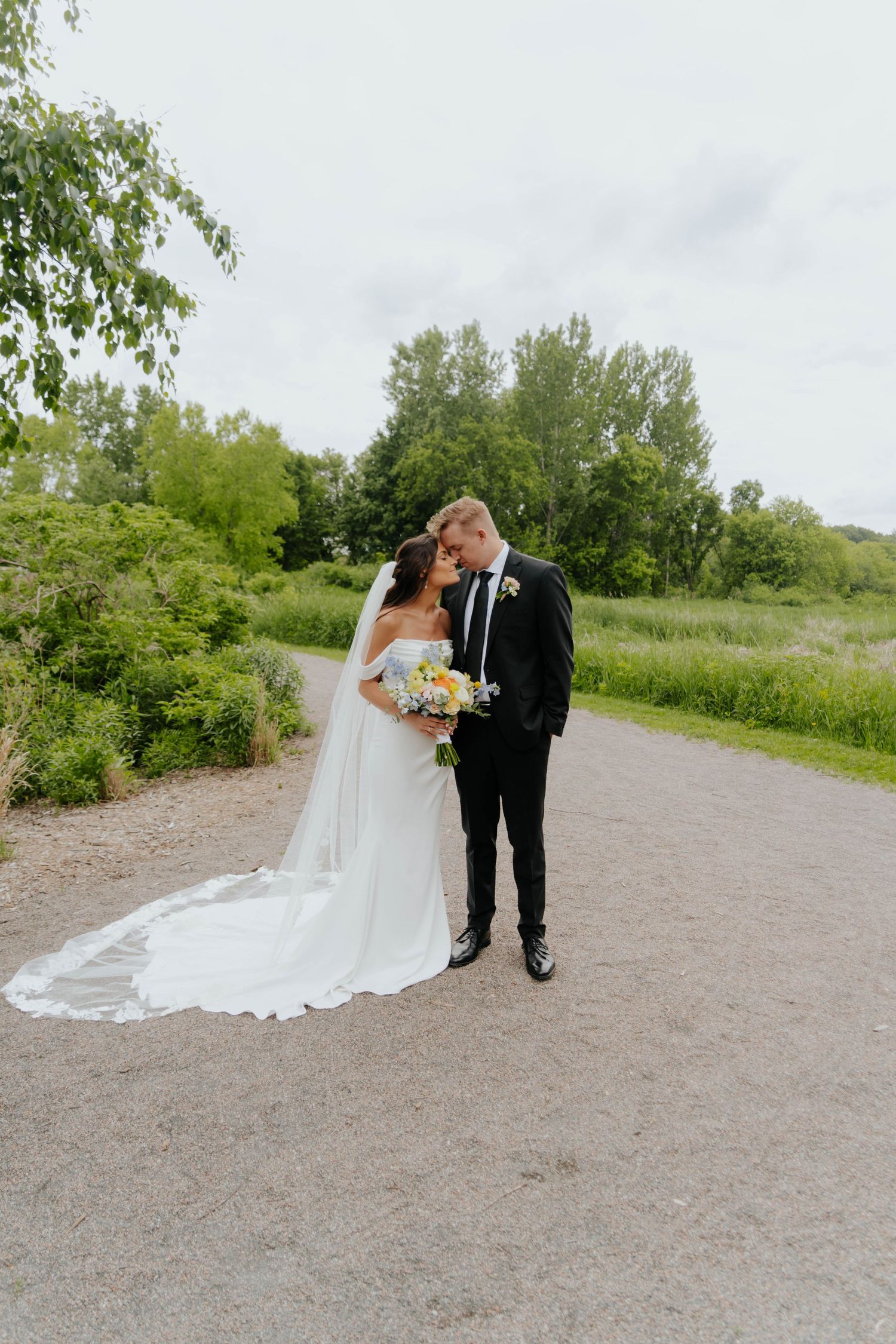 A bride and groom share an intimate moment on a dirt path surrounded by lush greenery on their wedding day.