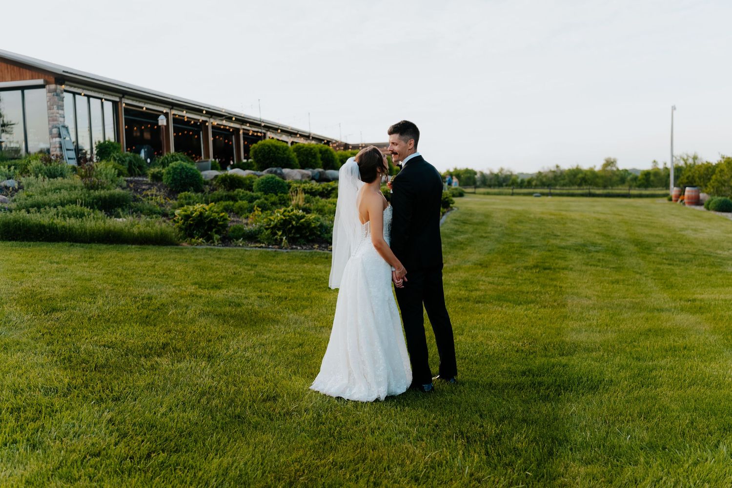 Bride and groom share a romantic kiss on lush green lawn outside elegant wedding venue at dusk.