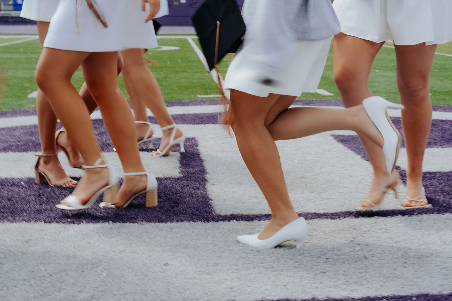 Women in white dresses and heels walking on a purple field during a graduation ceremony.