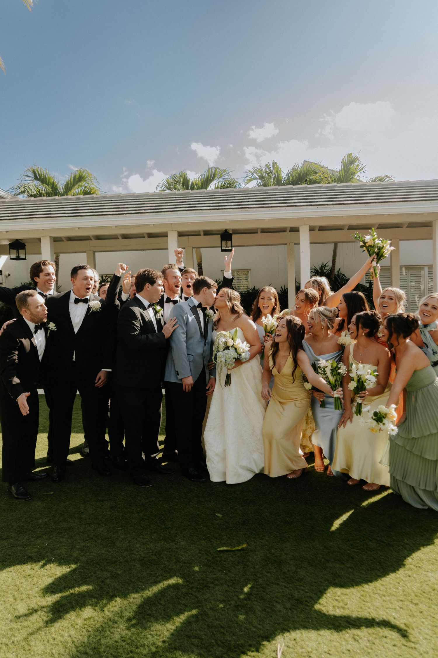Joyful wedding party celebrating outdoors on green lawn with bride, groom, groomsmen in tuxedos, and bridesmaids in sage gowns.