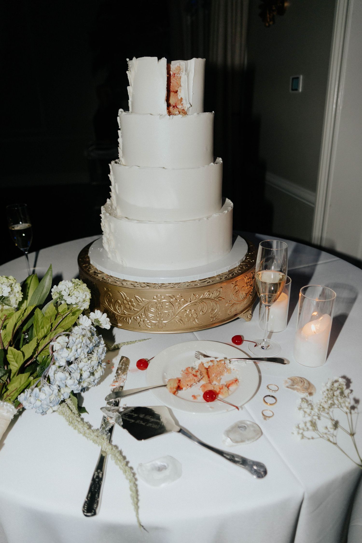 A four-tier white wedding cake on a gold stand, partially cut, displayed on a white tablecloth with champagne and candles.