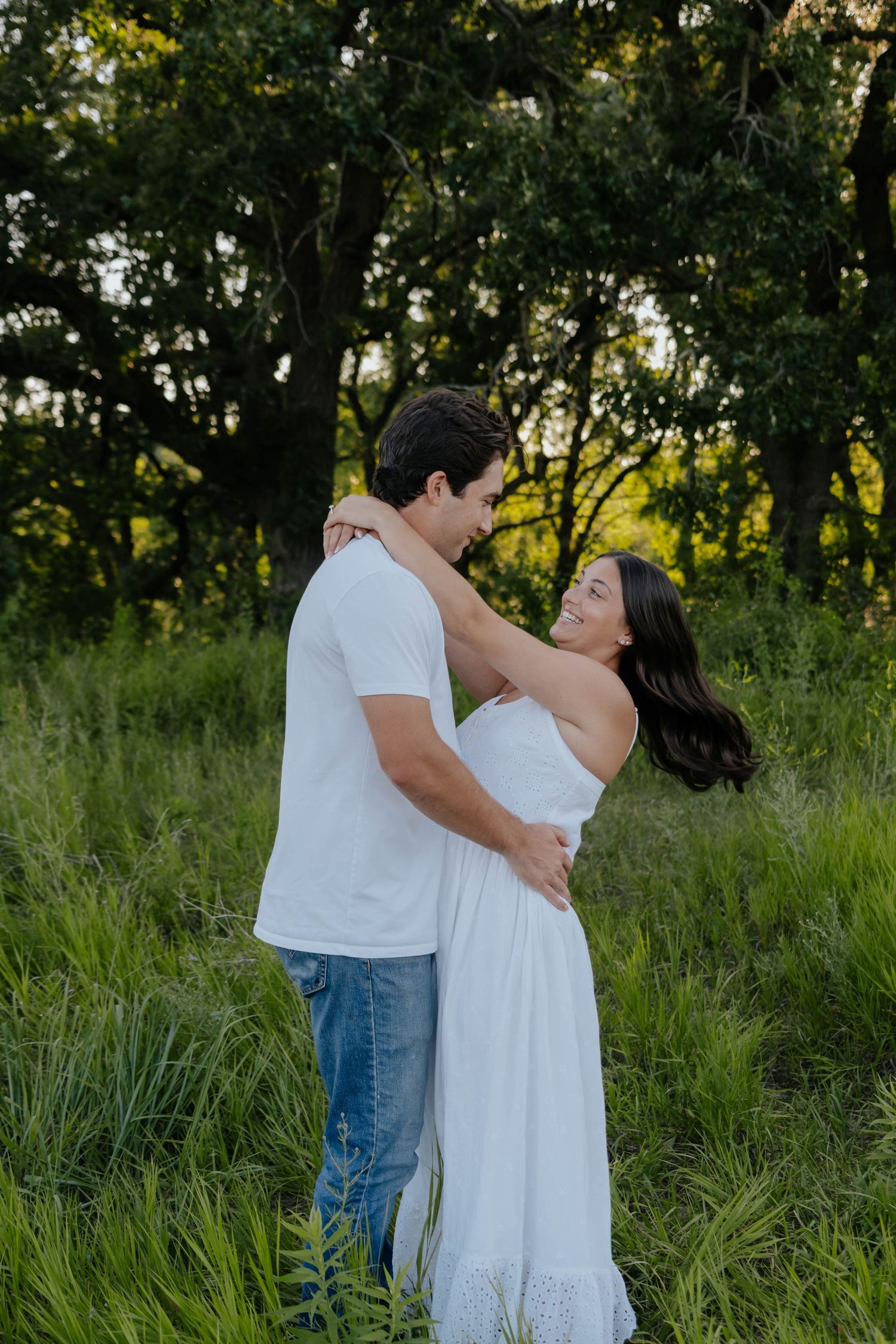 A romantic couple embraces in a sunlit meadow with tall grass and trees in the background.