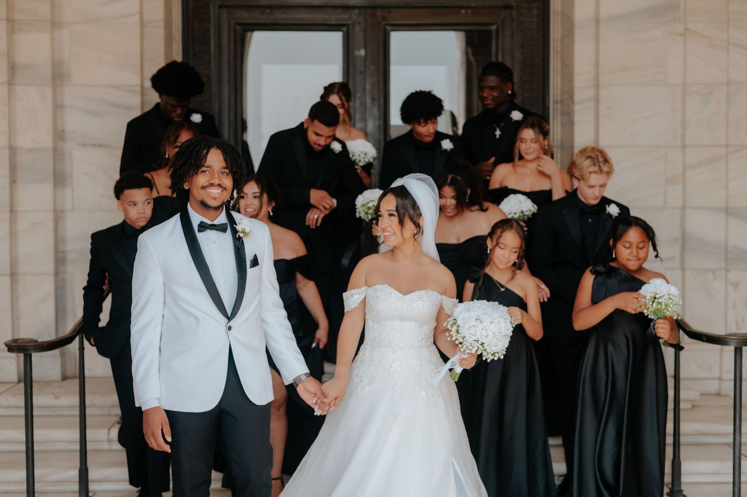 Bride and groom hold hands on stone steps with wedding party in black attire behind them at elegant venue.