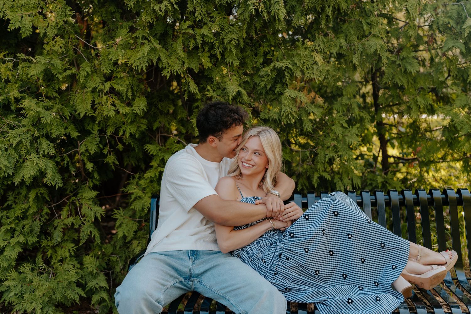 A couple sits closely on a park bench, smiling and embracing surrounded by lush green trees.