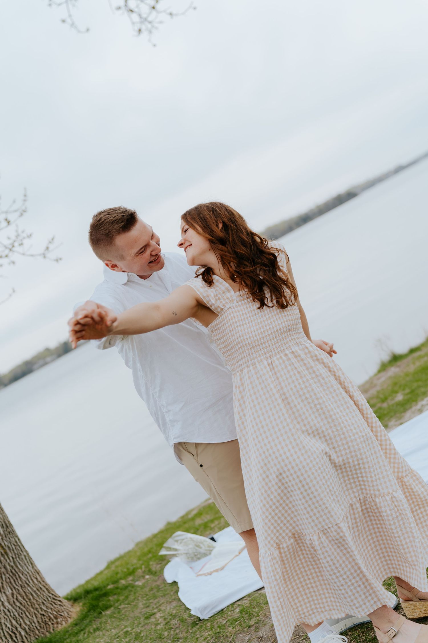 Happy couple dancing and holding hands by a lakeside, woman in a beige gingham dress, man in white shirt.