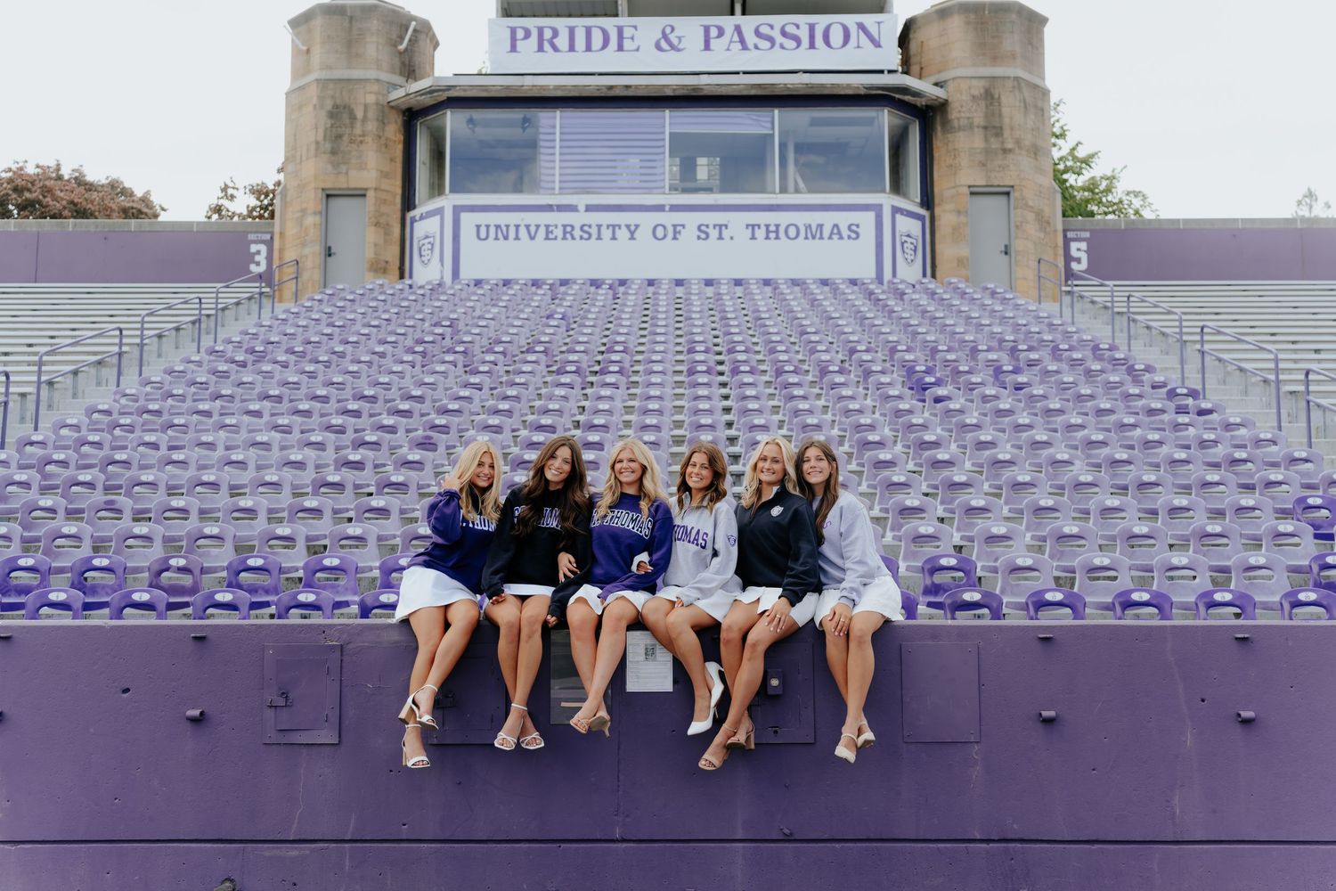 Six young women sit together on purple stadium bleachers at University of St. Thomas, smiling for a group photo.
