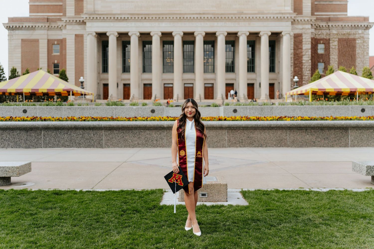 Graduate in cap and gown stole poses in front of a grand university building with fountains and yellow umbrellas.