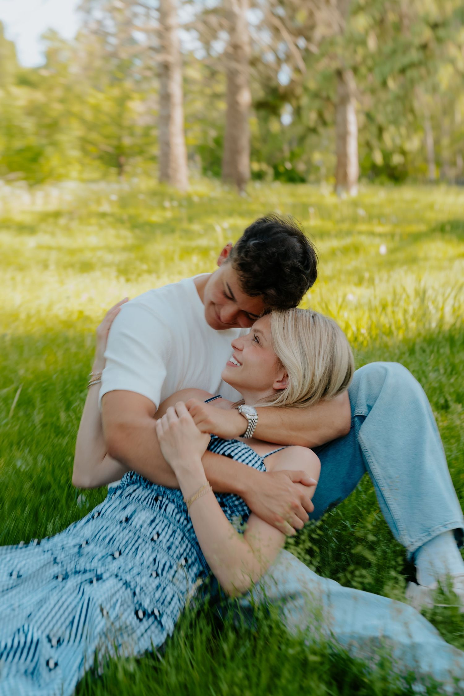Couple sitting on green grass in a park, woman leaning back on man as they share a tender romantic moment outdoors.