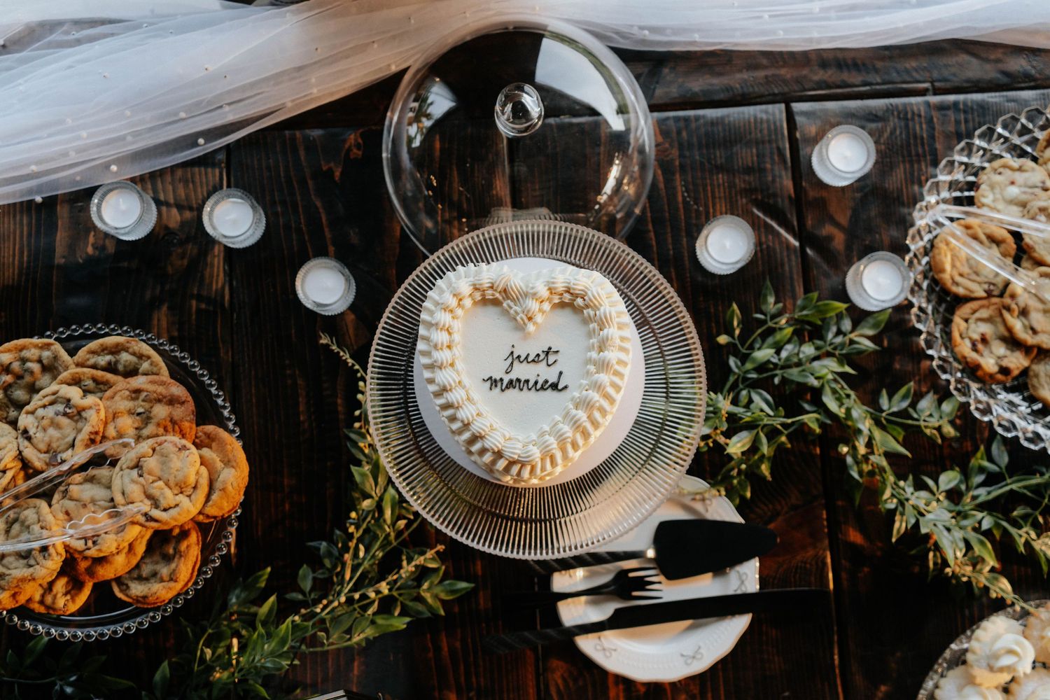 Heart-shaped wedding cake with 'just married' text surrounded by candles and pastries on a rustic wooden table.