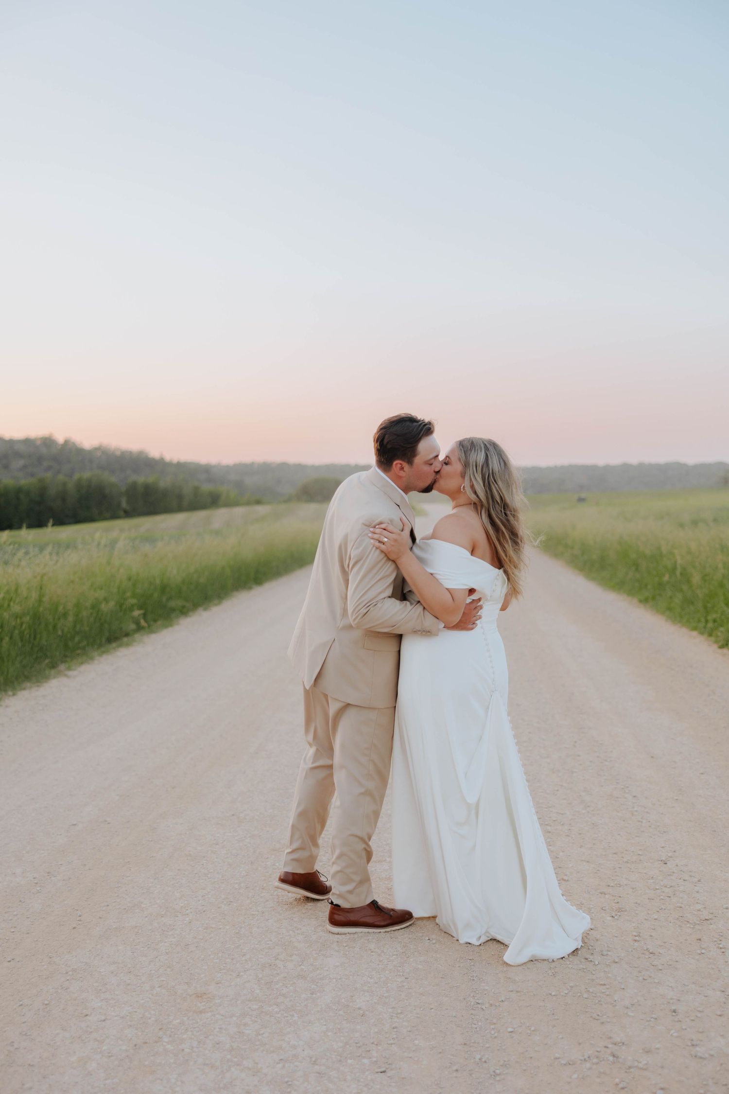 A couple shares a romantic kiss on a gravel road at sunset, bride in white gown and groom in tan suit.