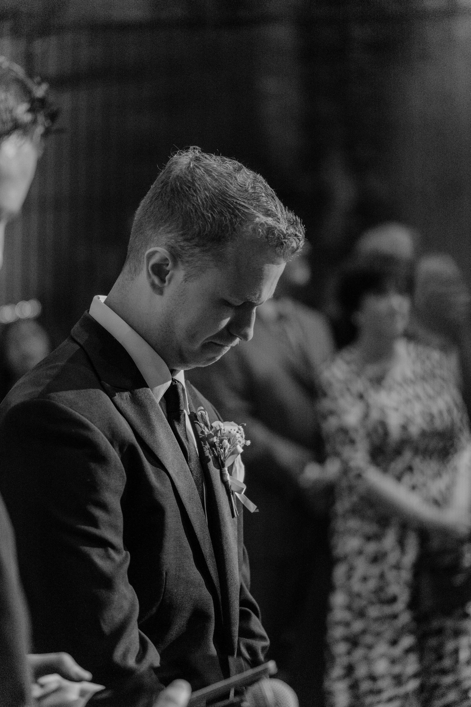Black and white portrait of a groom in a suit with boutonniere bowing his head during a wedding ceremony.