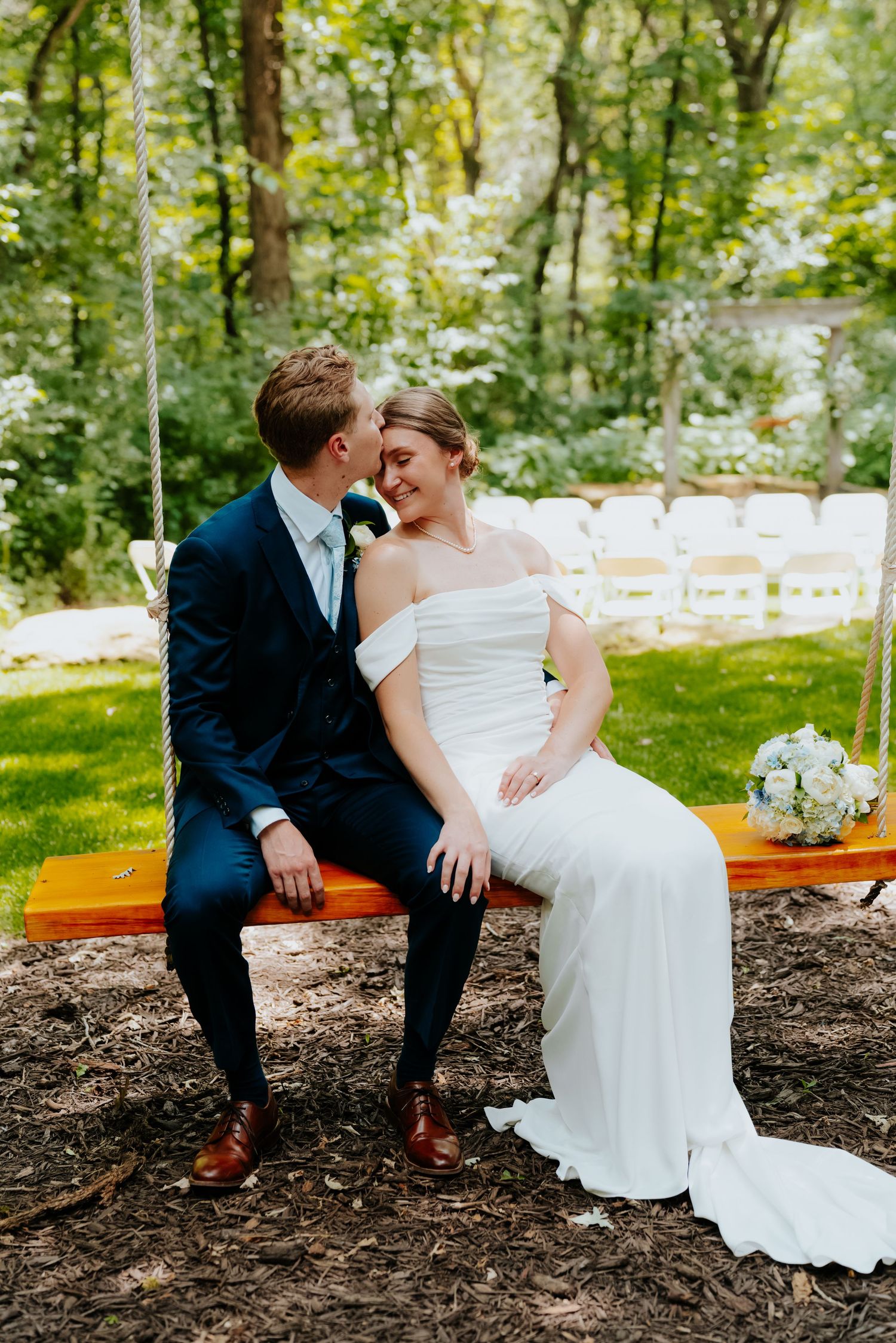 Bride and groom sitting on a wooden swing in a lush garden, smiling at each other on their wedding day.