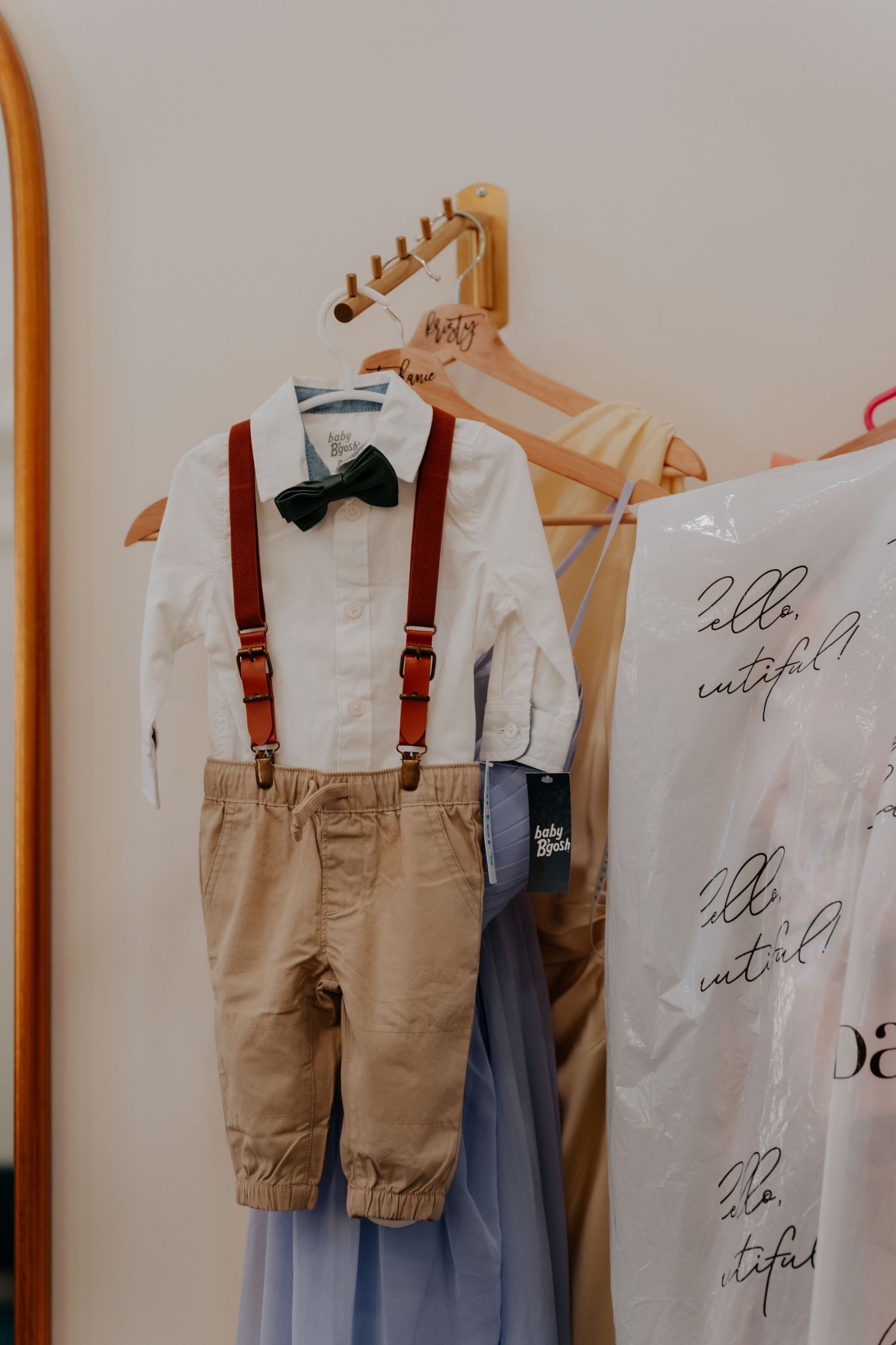 A little boys wedding outfit with white shirt, green bow tie, red suspenders, and khaki pants hanging on a wooden hanger.