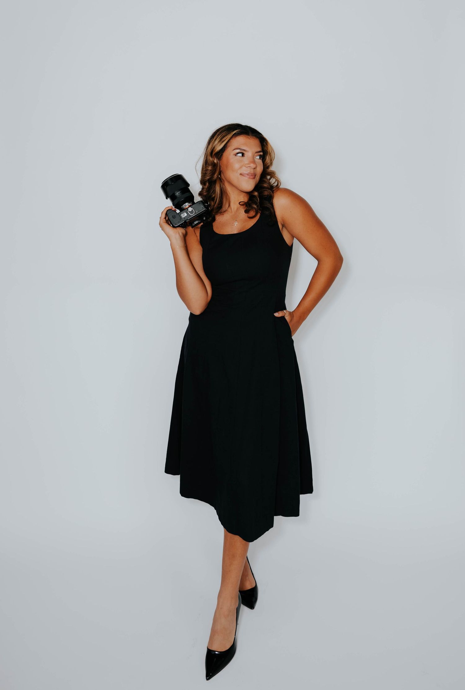 Woman in black dress holding camera, posing confidently against light gray background, wearing black heels.